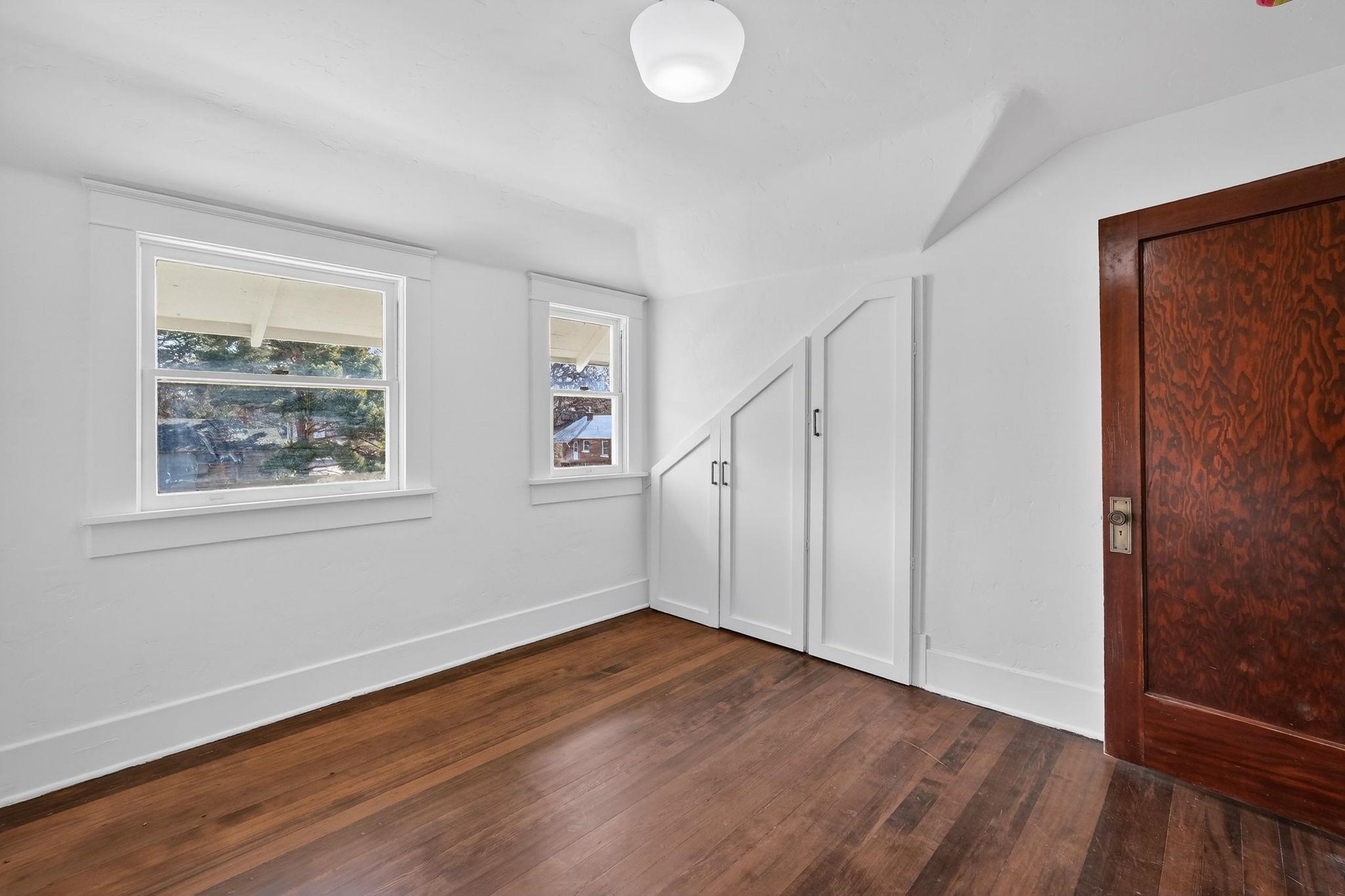 Empty room with hardwood floors, two windows, and built-in cabinets beneath a sloped ceiling.