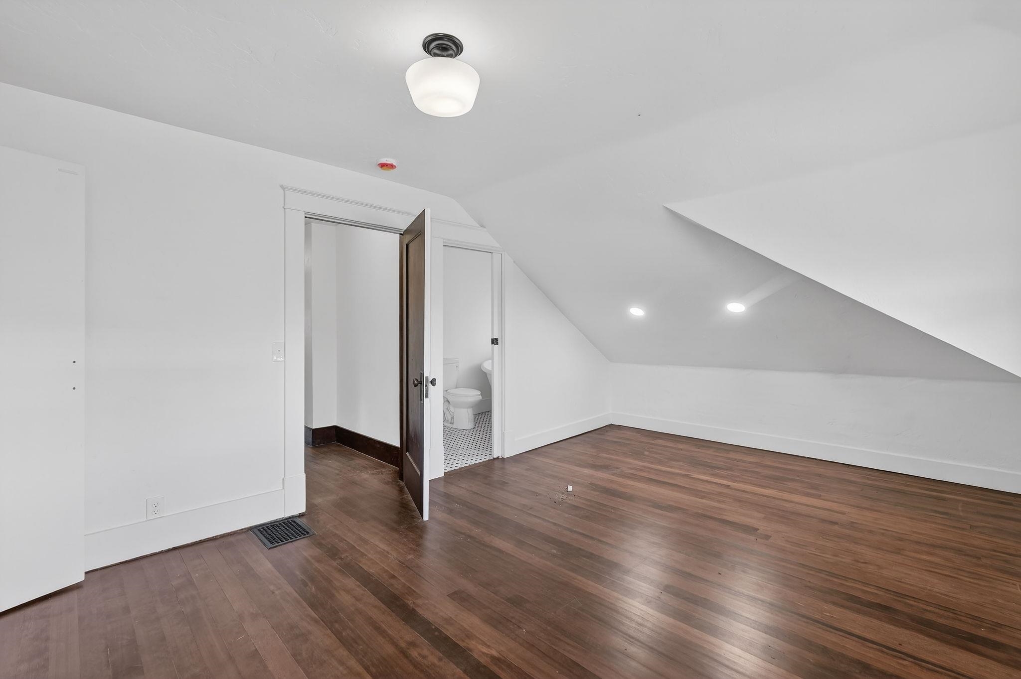 Empty attic room with dark wood floors and a bathroom visible through the open doorway.
