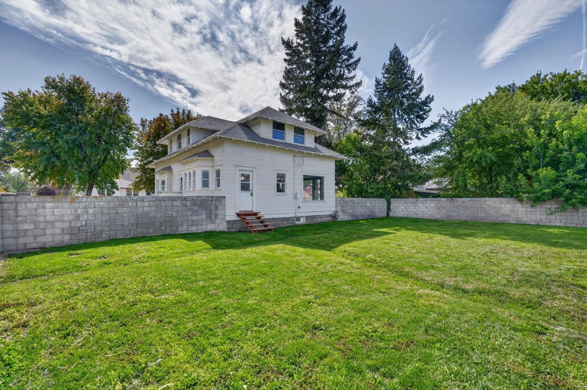 White two-story house beyond a spacious green backyard enclosed by a concrete block wall.