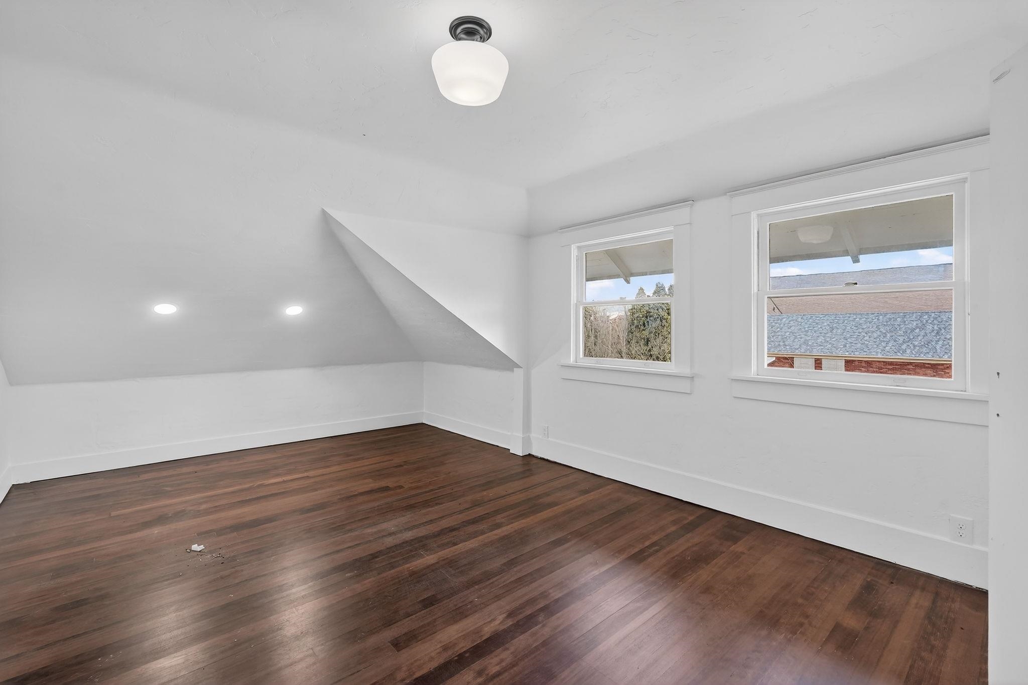 Vacant upper-floor room with sloped ceilings, dark hardwood floors, and two windows.