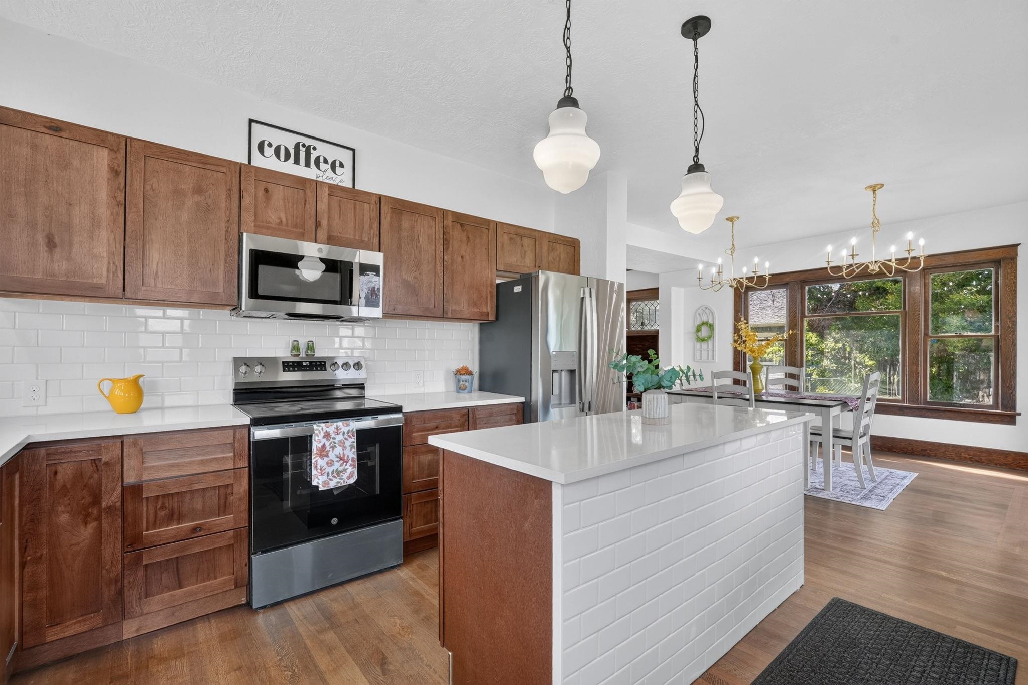 Kitchen with a large tiled island, warm wood cabinetry, stainless appliances, and an adjoining dining area by broad windows.