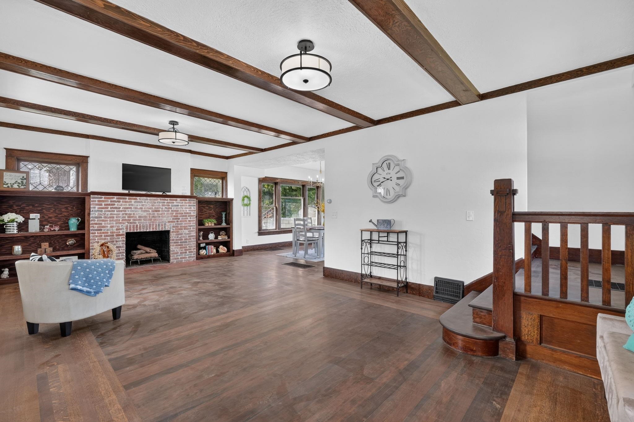 Living room with exposed wood ceiling beams, brick fireplace, built-in shelves, and hardwood floors.