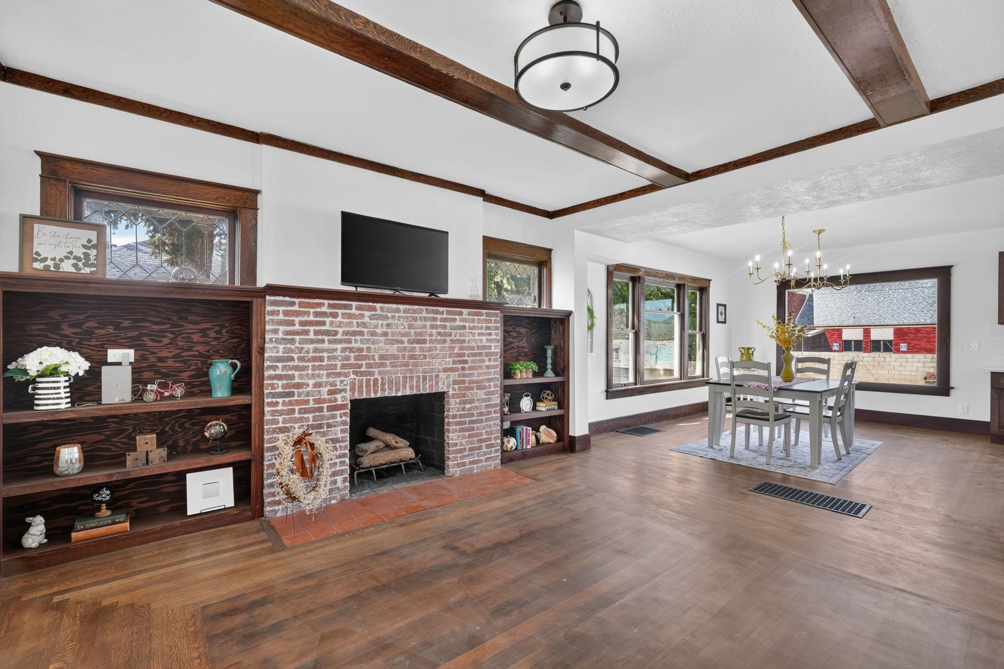 Living room with a brick fireplace, dark built-in shelves, exposed ceiling beams, and an adjoining dining area.