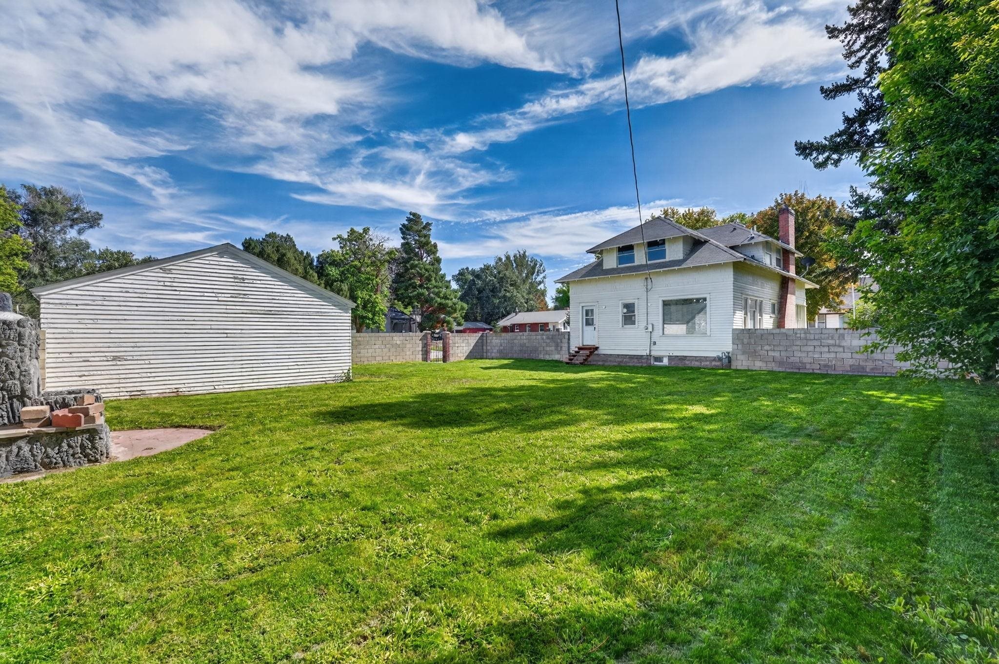 White two-story house and detached garage beyond a broad lawn and stone outdoor fireplace.