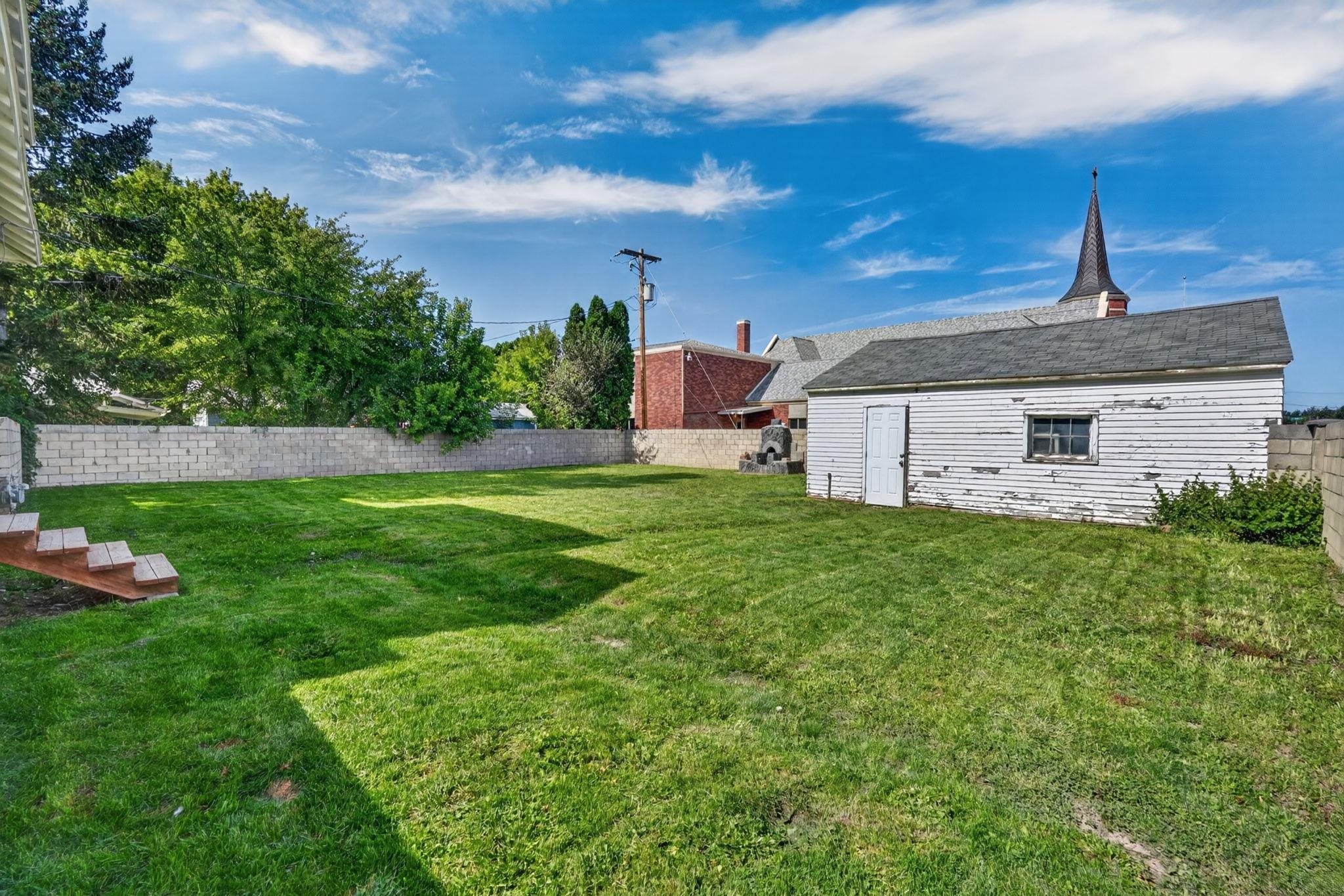 Green backyard with a weathered white outbuilding, wooden steps, and a masonry boundary wall.
