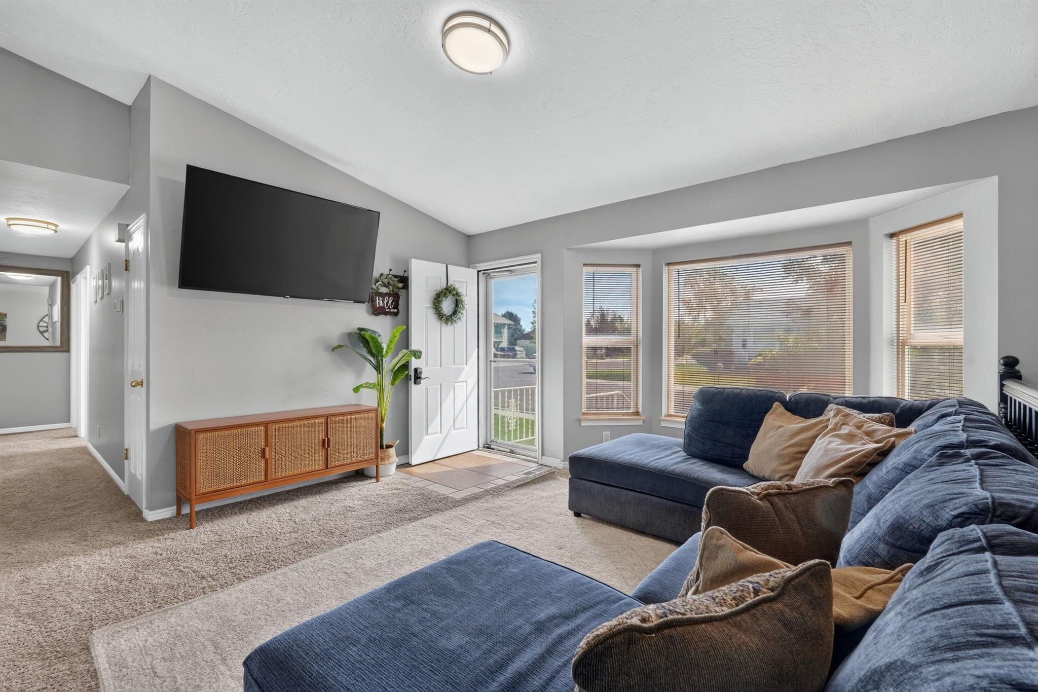 Living room with a blue sectional, wall-mounted TV, and open front door beside bright windows.