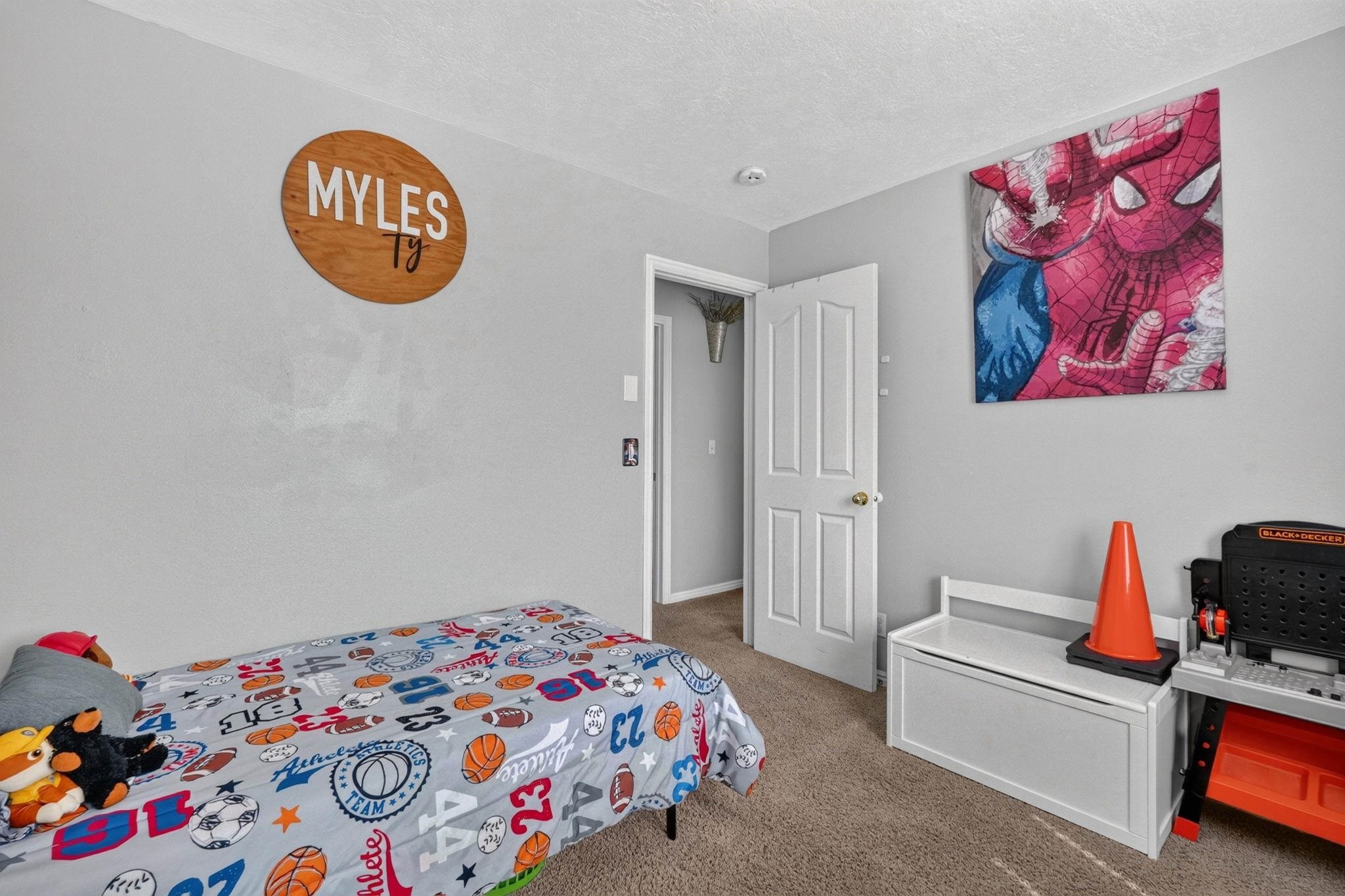 Child’s bedroom with sports-print bedding, a wooden name sign, Spider-Man artwork, and a white storage bench.