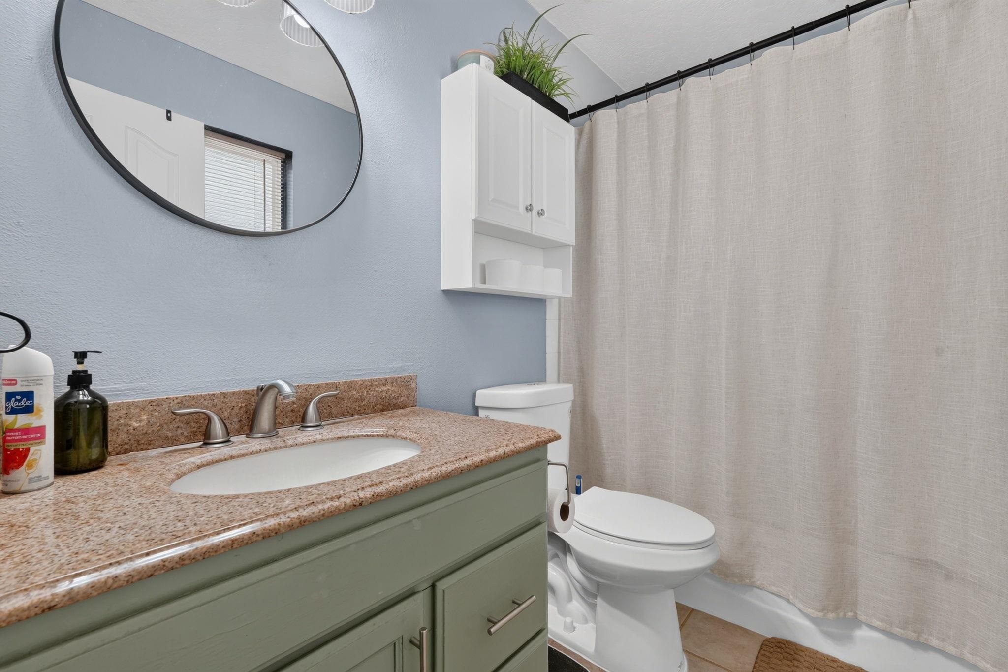 Bathroom with a sage-green vanity, speckled countertop, round mirror, and beige shower curtain.
