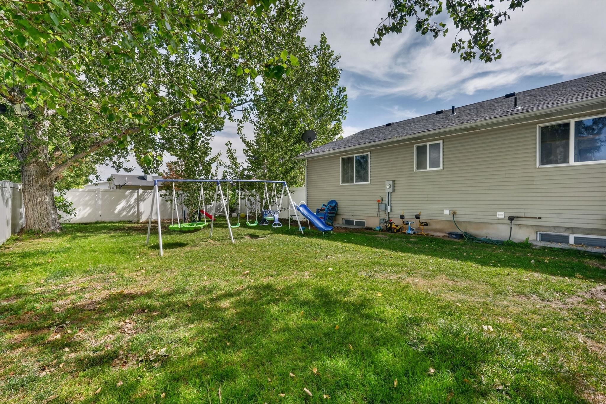 Backyard with a grassy lawn, mature trees, and a swing set with a blue slide beside the house.