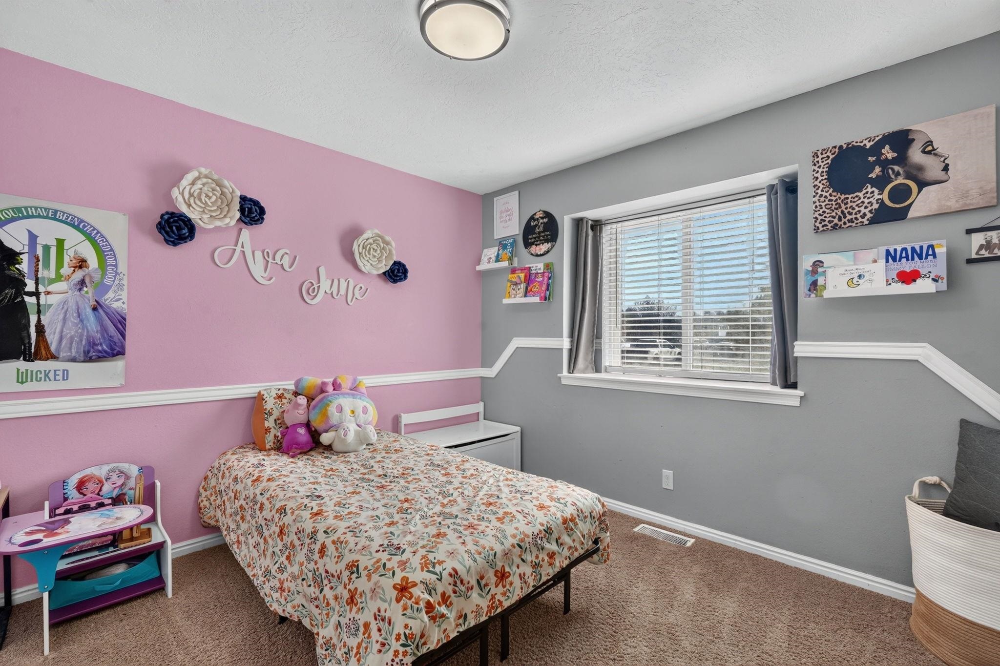 Bedroom with a pink accent wall, floral bedspread, stuffed toys, and window with blinds.