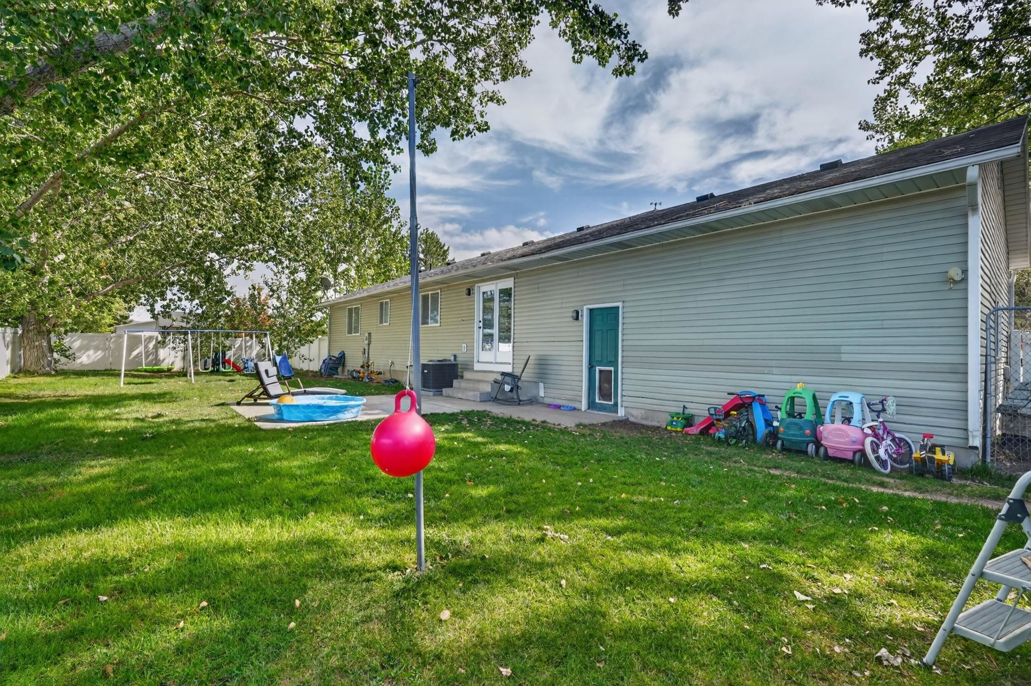 Home’s backyard with a small pool, swing set, mature trees, and children’s toys.
