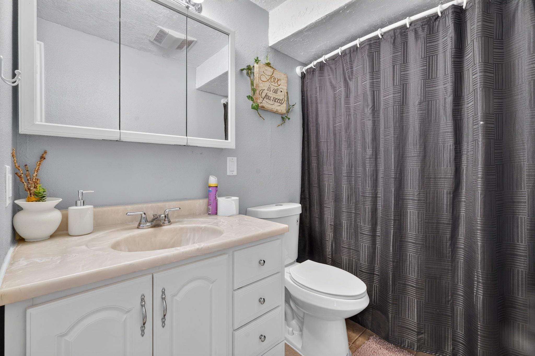 Bathroom with a white vanity, beige countertop, toilet, and dark patterned shower curtain.
