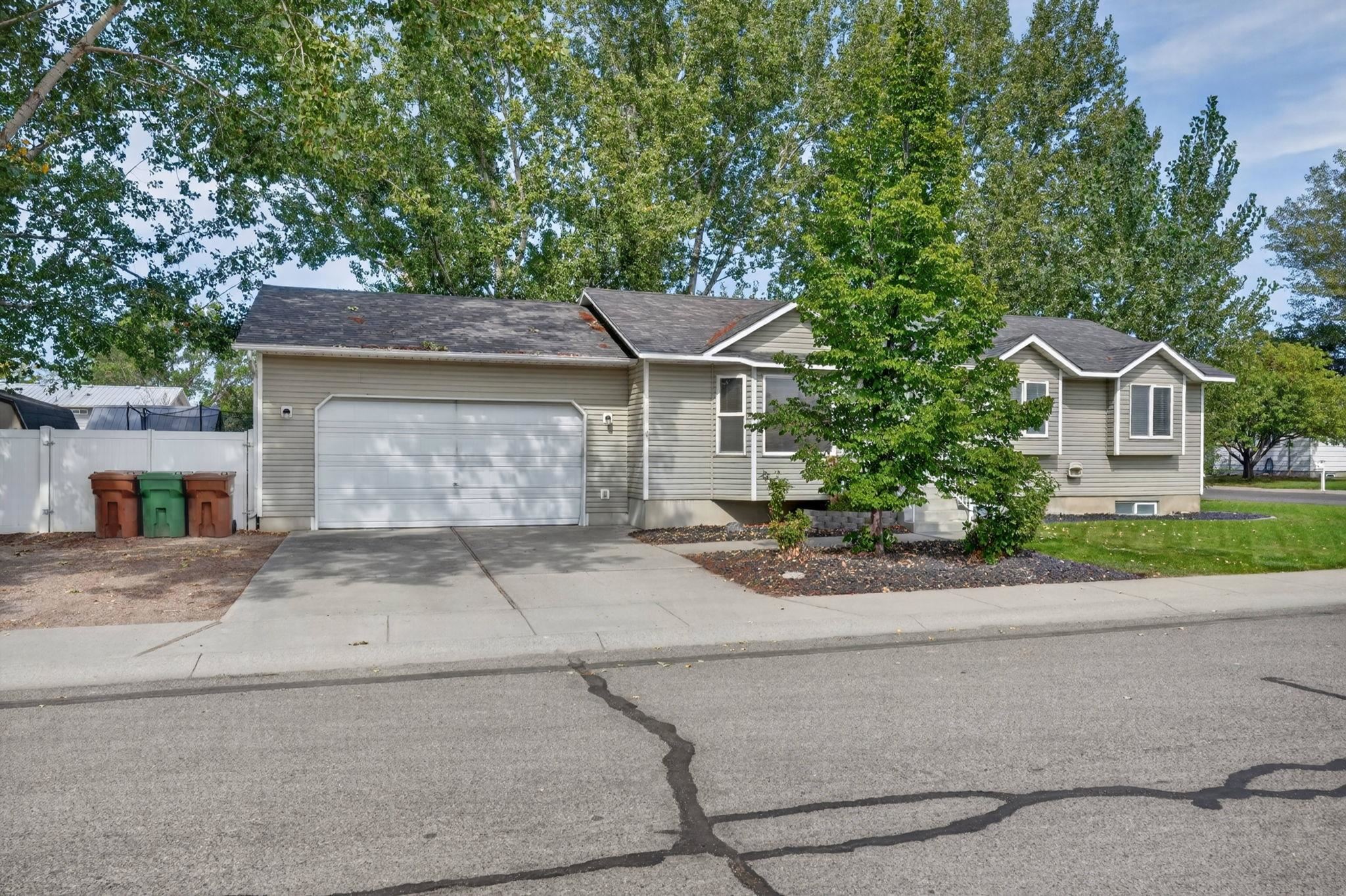 Gray-sided house with a two-car garage, concrete driveway, mature trees, and a small front lawn.