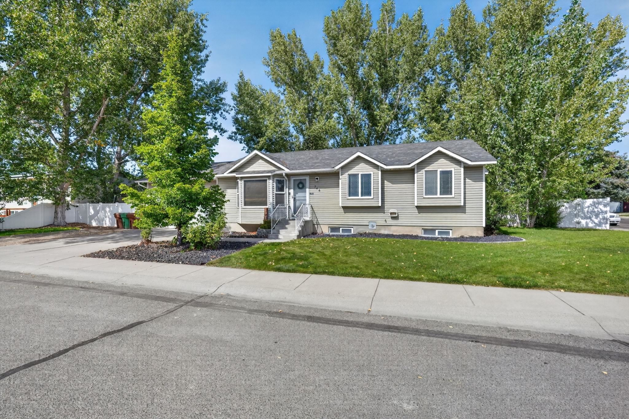 Gray-sided single-story home with a raised front entry, green lawn, mature trees, and driveway.