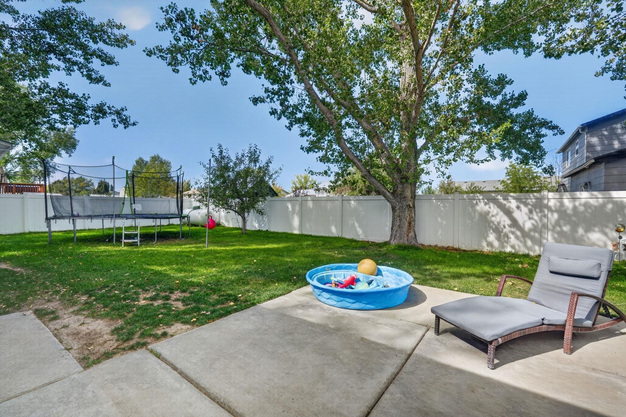 Fenced backyard with a large shade tree, trampoline, and patio with a lounge chair and blue splash pool.