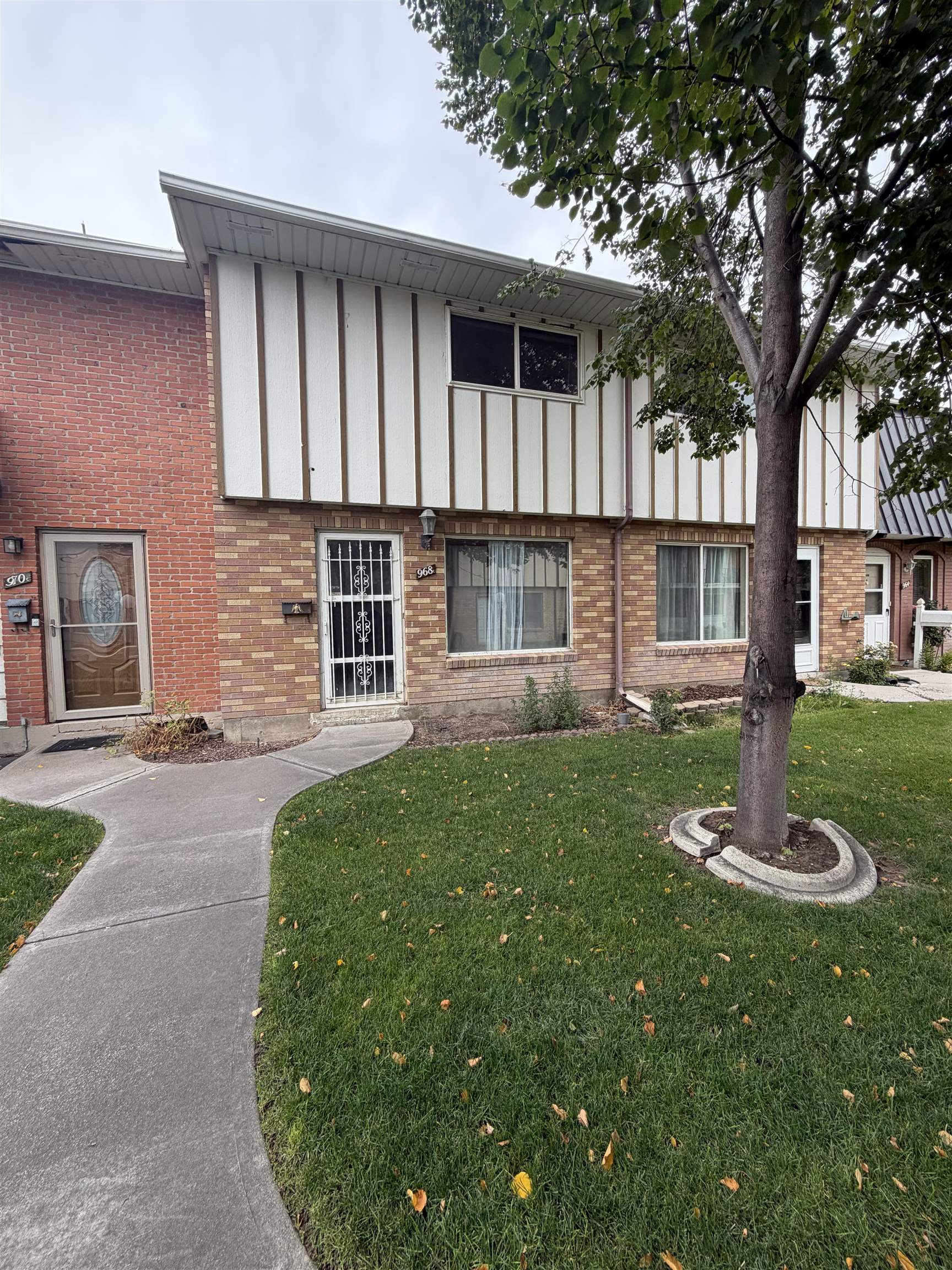 Two-story brick townhouse with white upper siding, a barred entry door, and a large tree in the lawn.