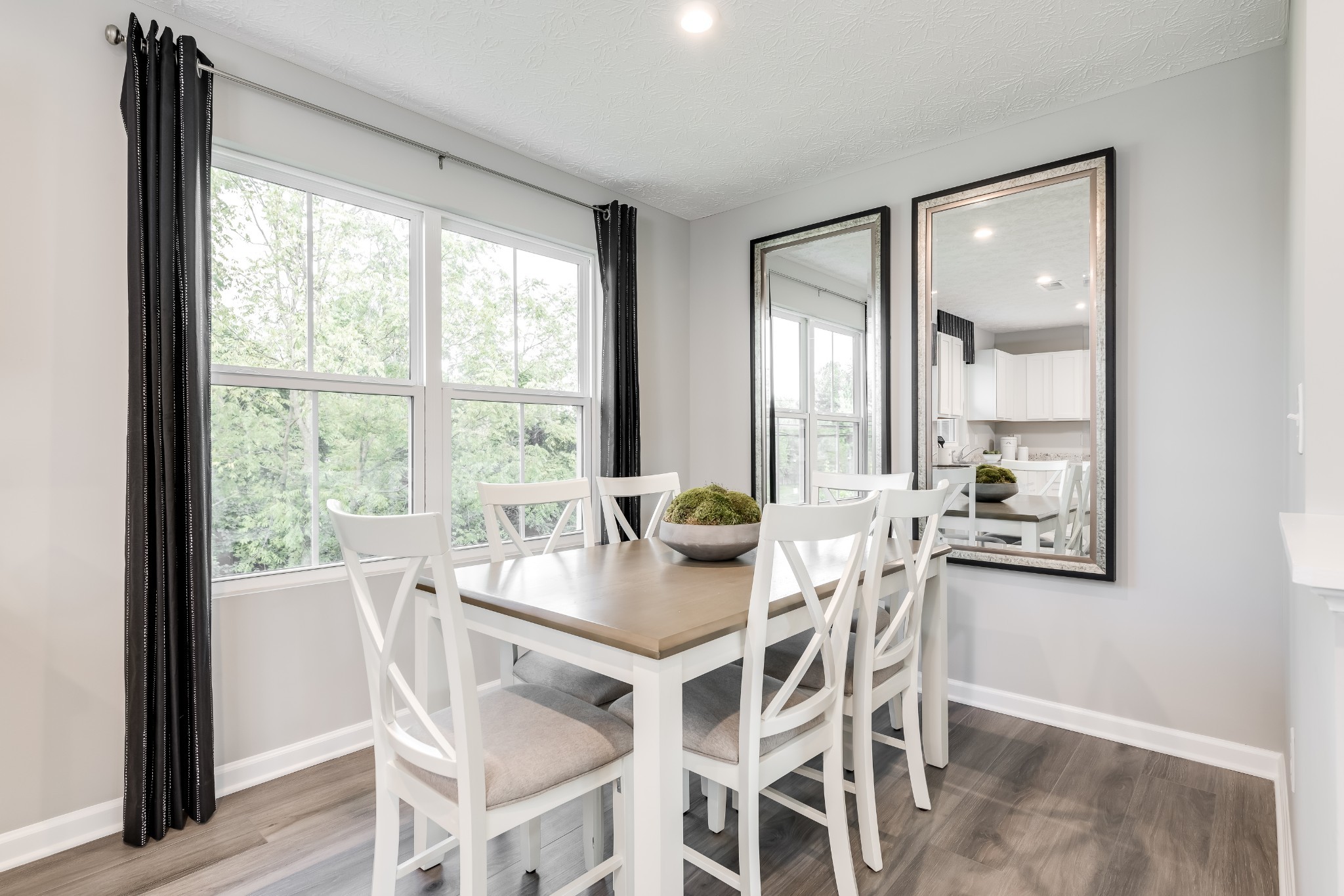 Dining room with a light wood table and white chairs beside large windows and a mirror reflecting the kitchen.