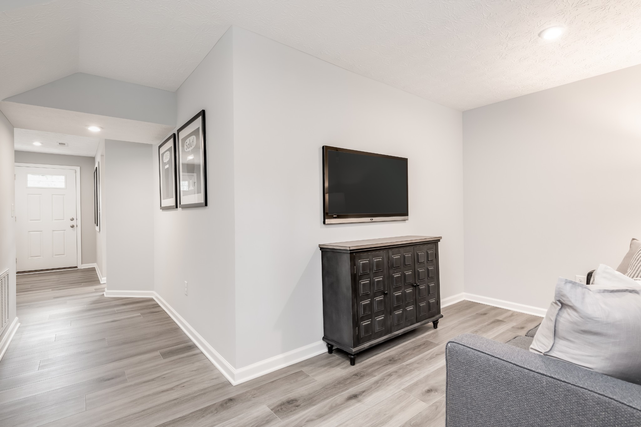 Living room with a wall-mounted television, dark console, gray sofa, and entry hallway.