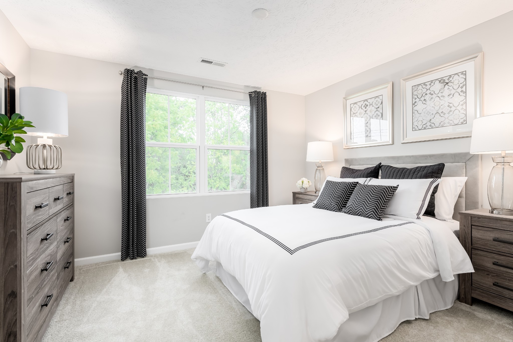 Staged bedroom with a white bed, black patterned accents, gray dressers, and a large window overlooking trees.