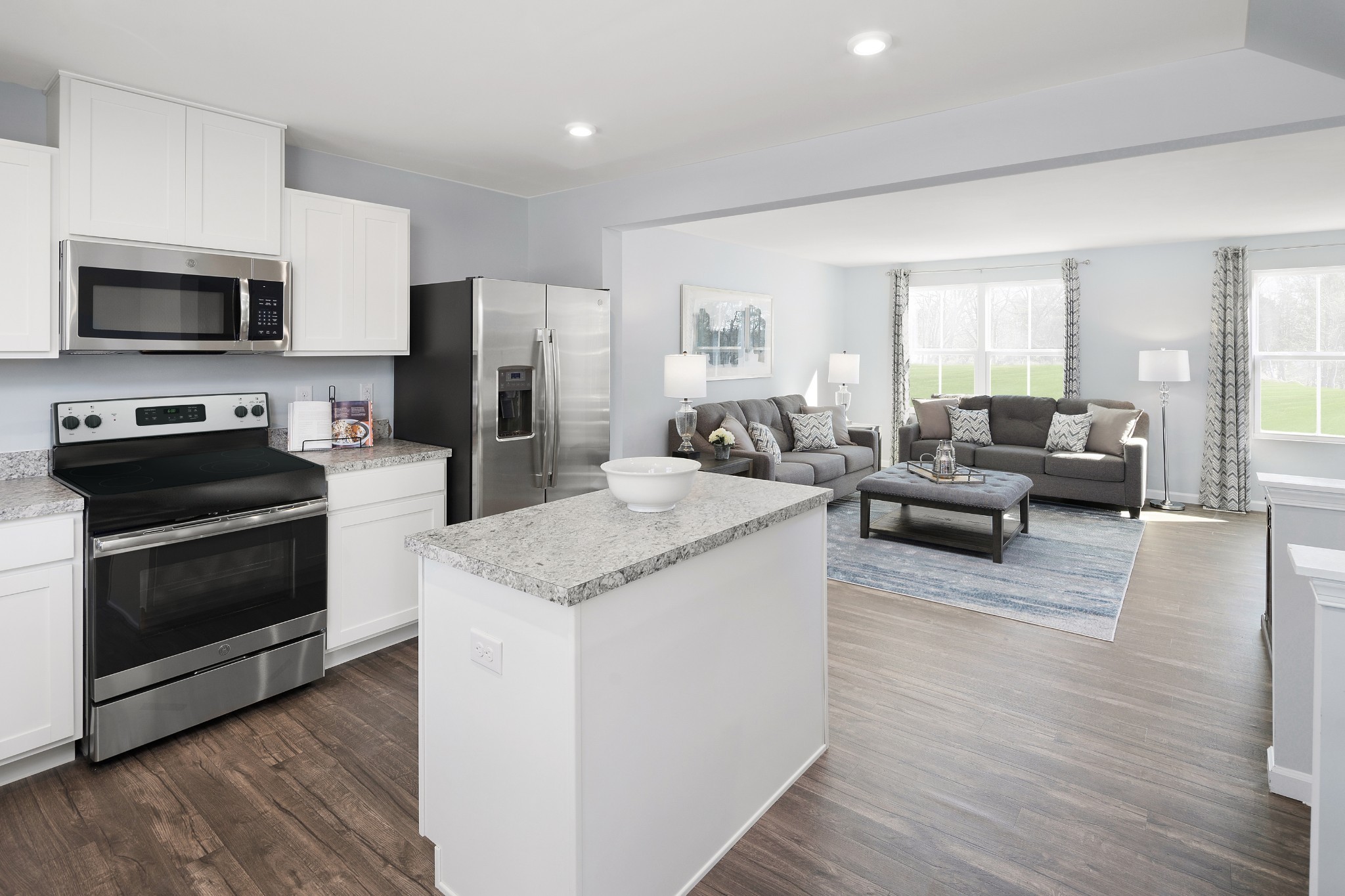 Open-plan kitchen and living room with a granite island, stainless appliances, and gray sofas by large windows.