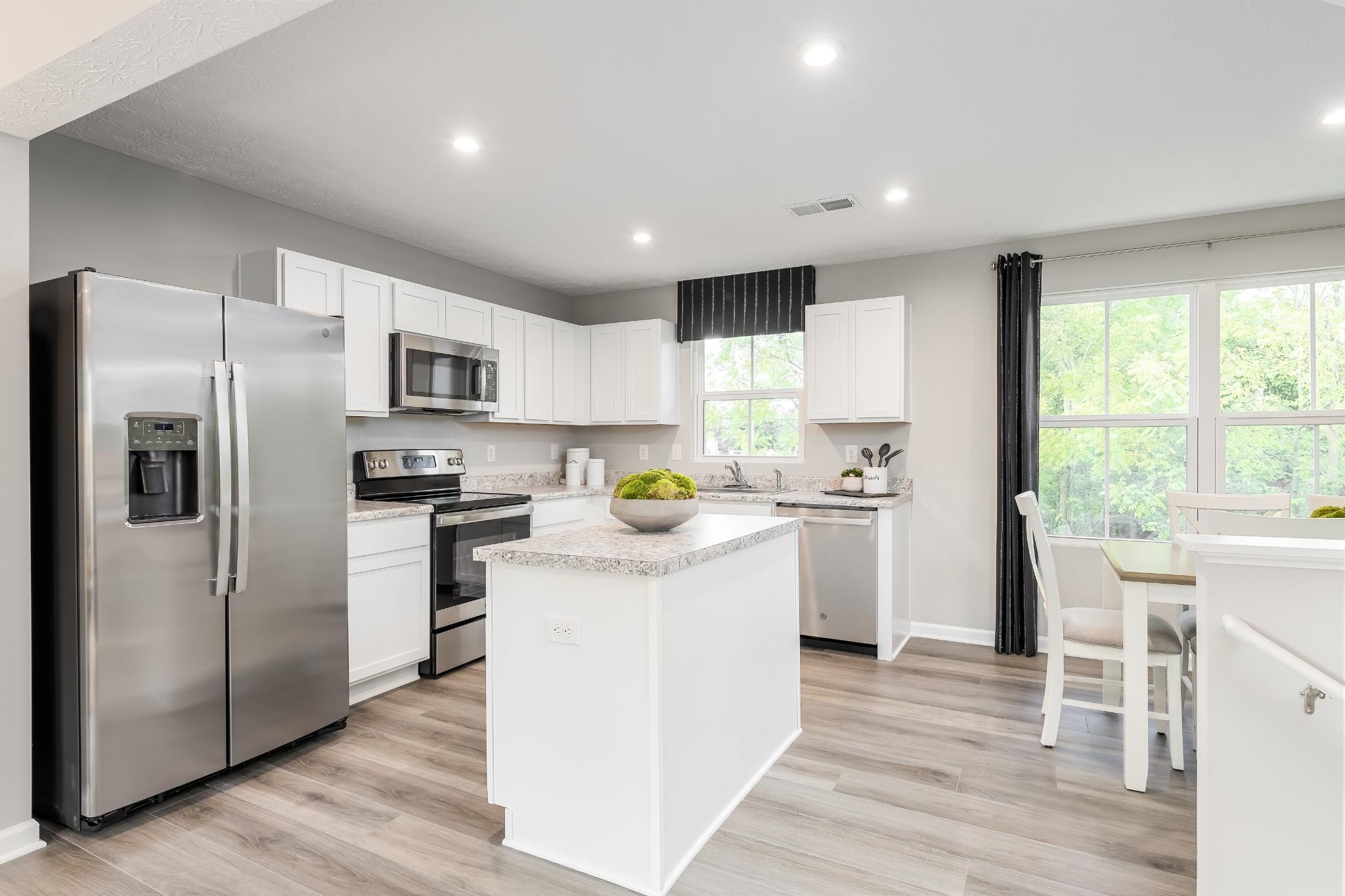 Bright kitchen with white cabinets, stainless steel appliances, and a central island beside a window-lit dining area.