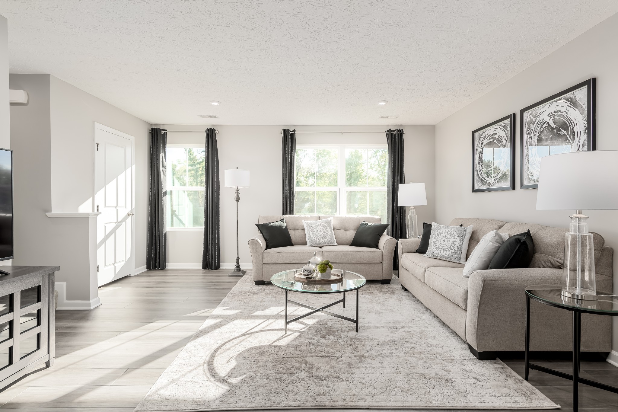 Staged living room with beige sofas, a glass coffee table, and sunlight streaming through large windows.