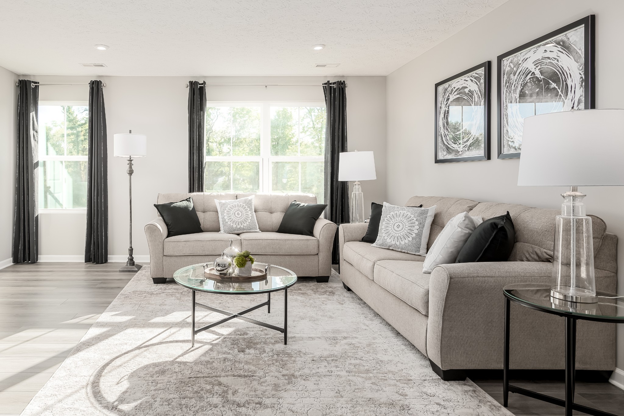 Staged living room with two beige sofas, a glass coffee table, and large sunlit windows.