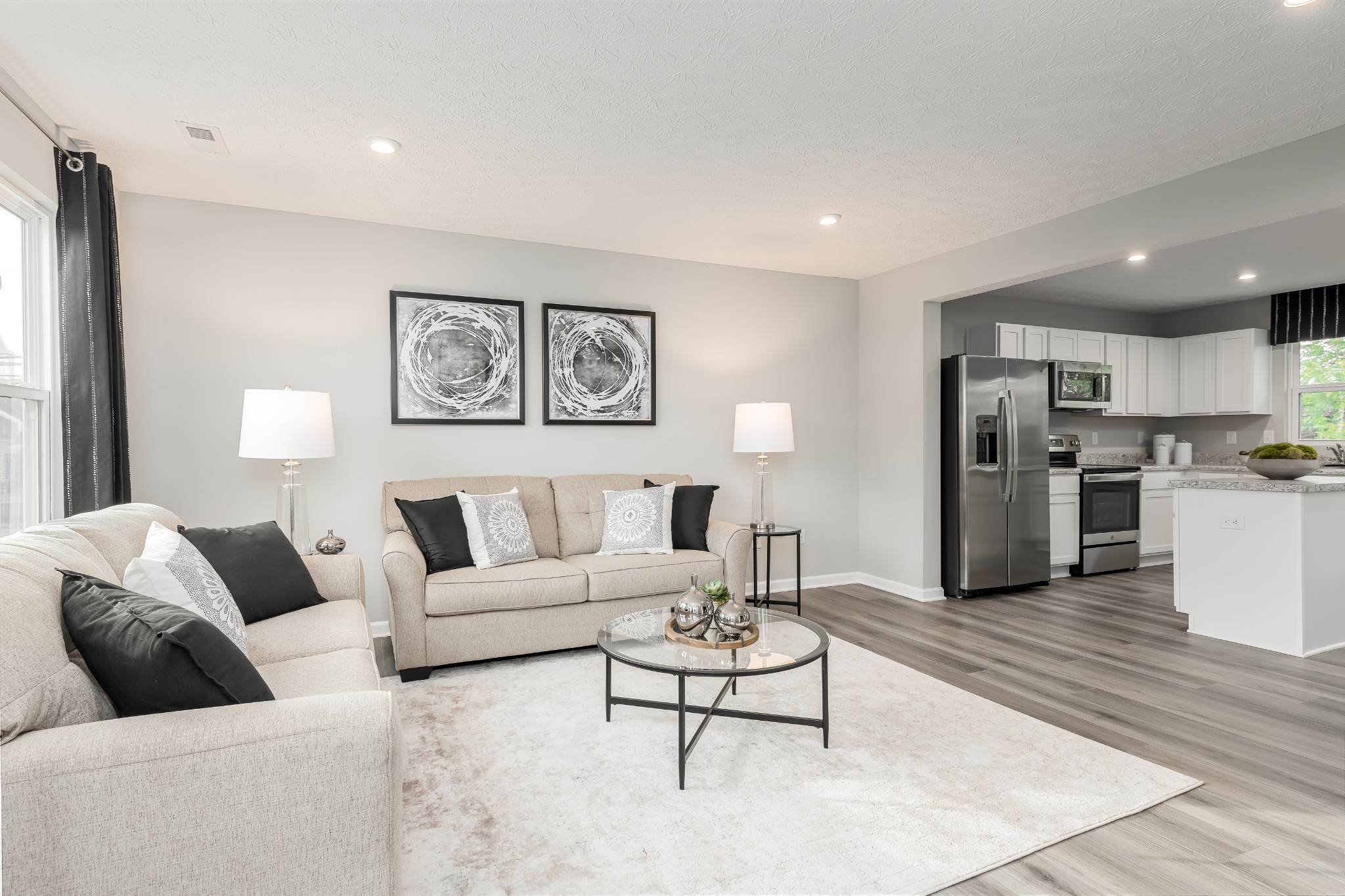 Staged living room opens to a white kitchen with stainless steel appliances and gray flooring.