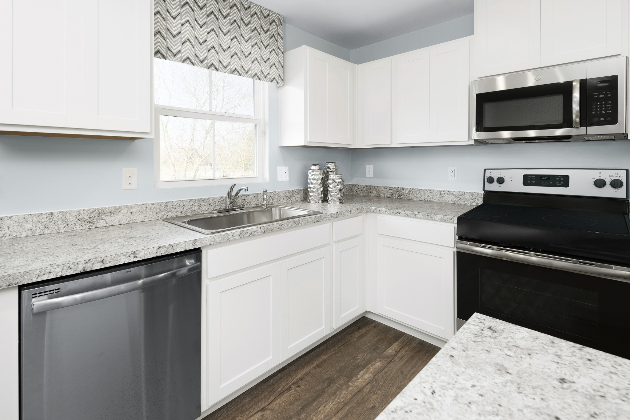 Kitchen with white cabinets, speckled stone counters, stainless appliances, and a chevron shade above the sink.