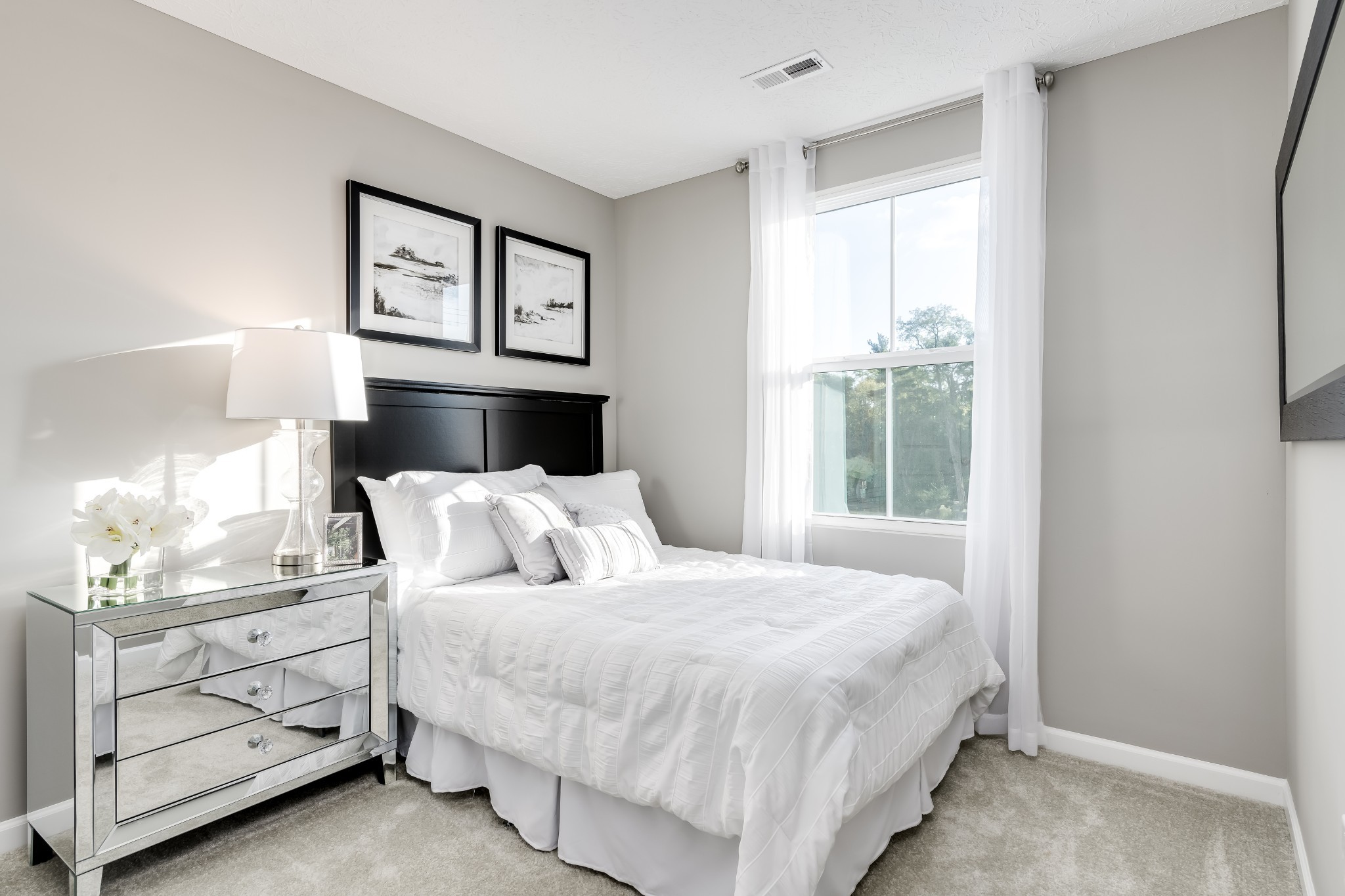 Bright bedroom with a white bed, black headboard, mirrored dresser, and large curtained window.