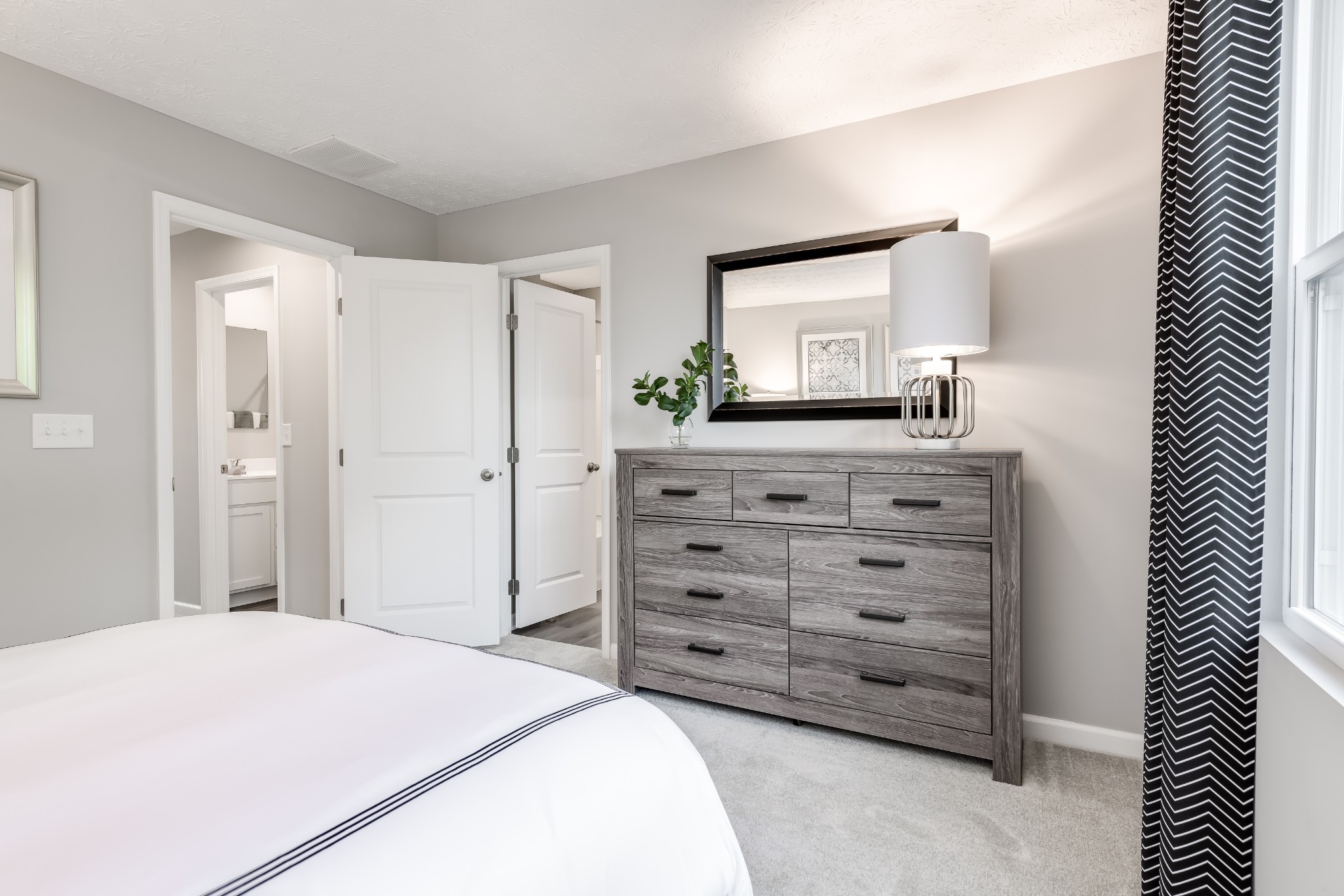 Staged bedroom with a gray dresser and mirror beside an open bathroom doorway.