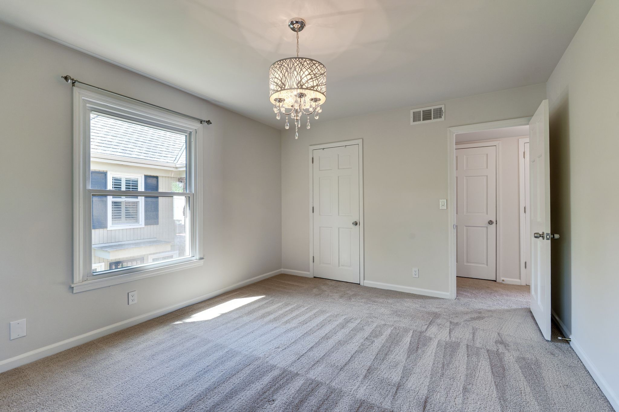 Empty carpeted bedroom with a crystal chandelier, large window, and open doorway.