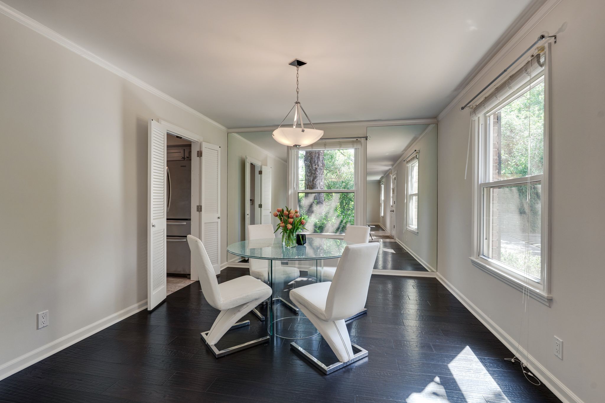 Dining room with a glass table, white chairs, dark floors, and large windows.