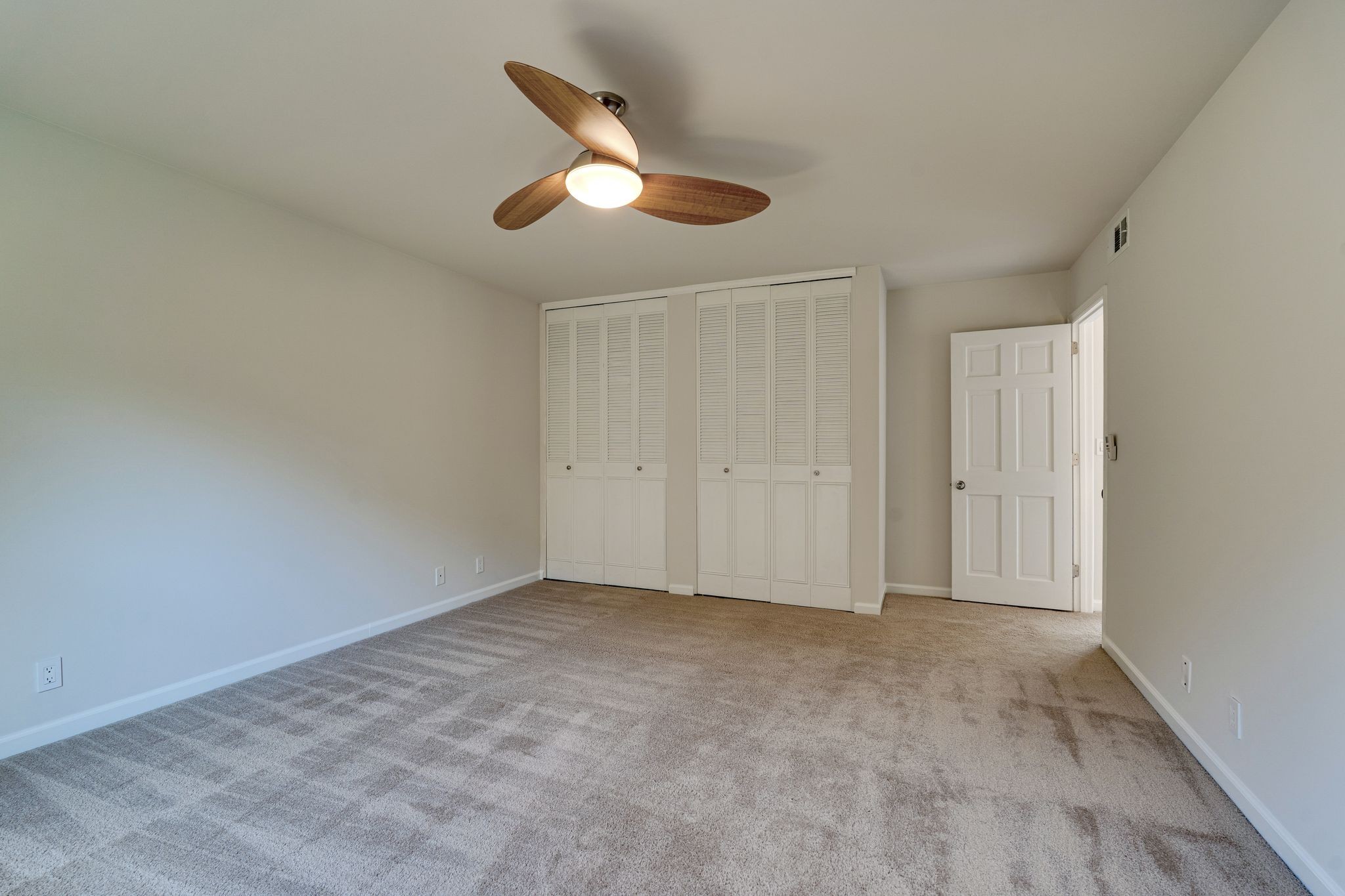 Empty bedroom with beige carpet, louvered closets, and a wood-blade ceiling fan.