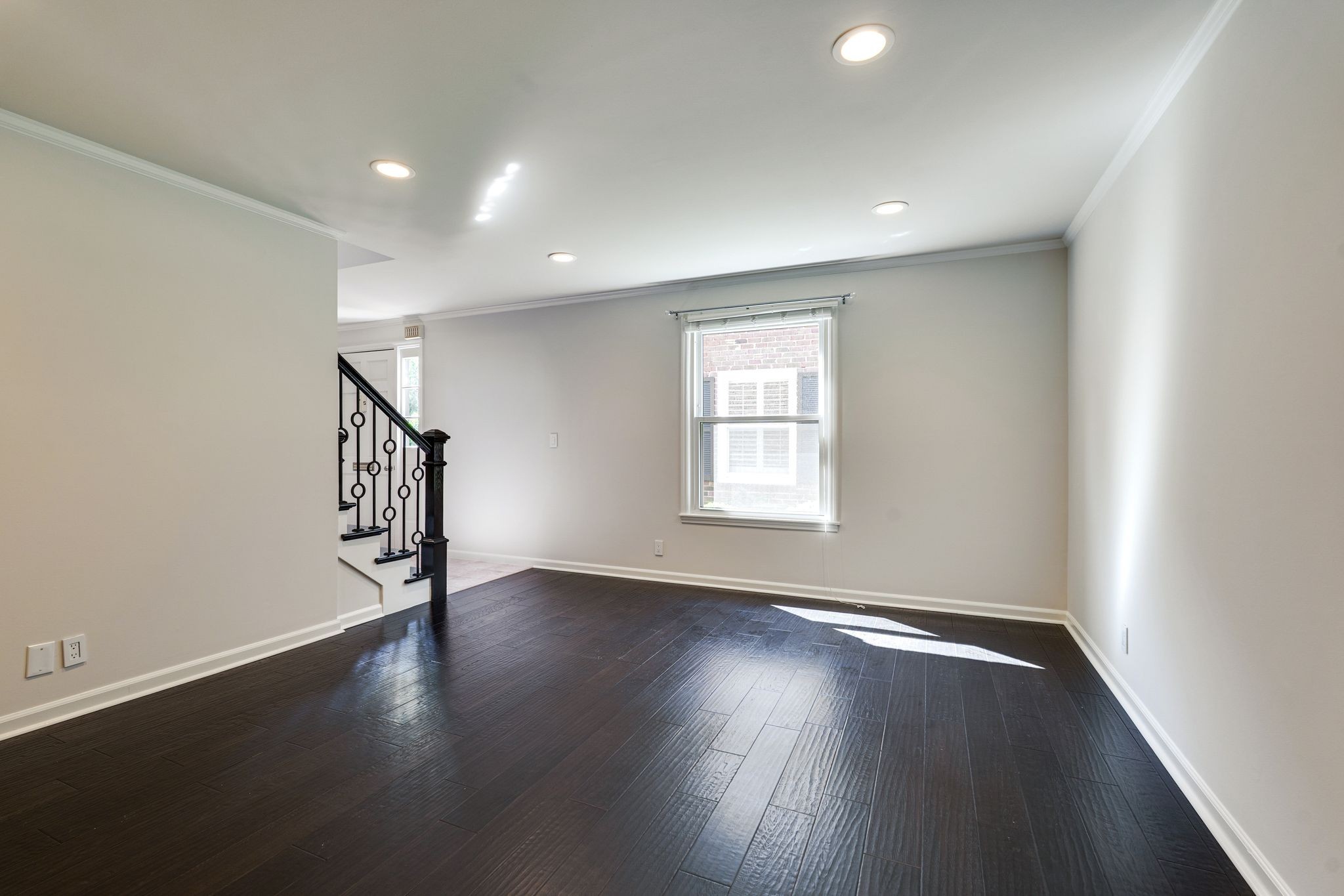 Empty room with dark wood floors, a sunlit window, and a black metal staircase railing.