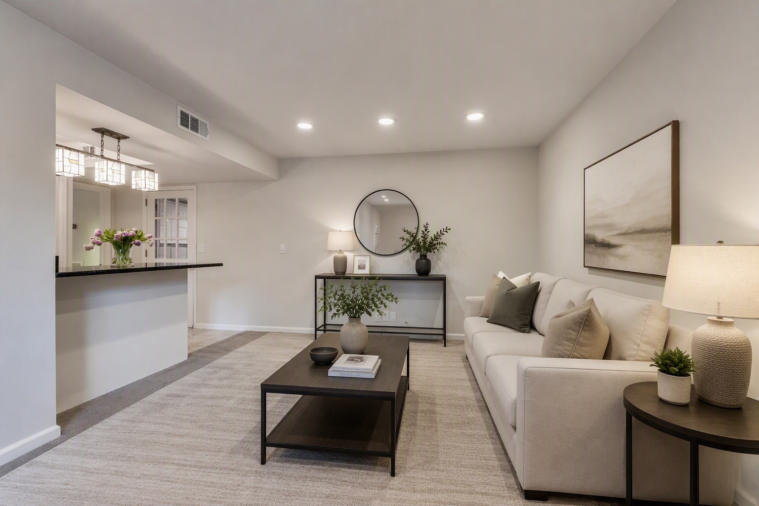Staged living room with a white sectional sofa, neutral rug, and dark coffee table beside a pass-through counter.