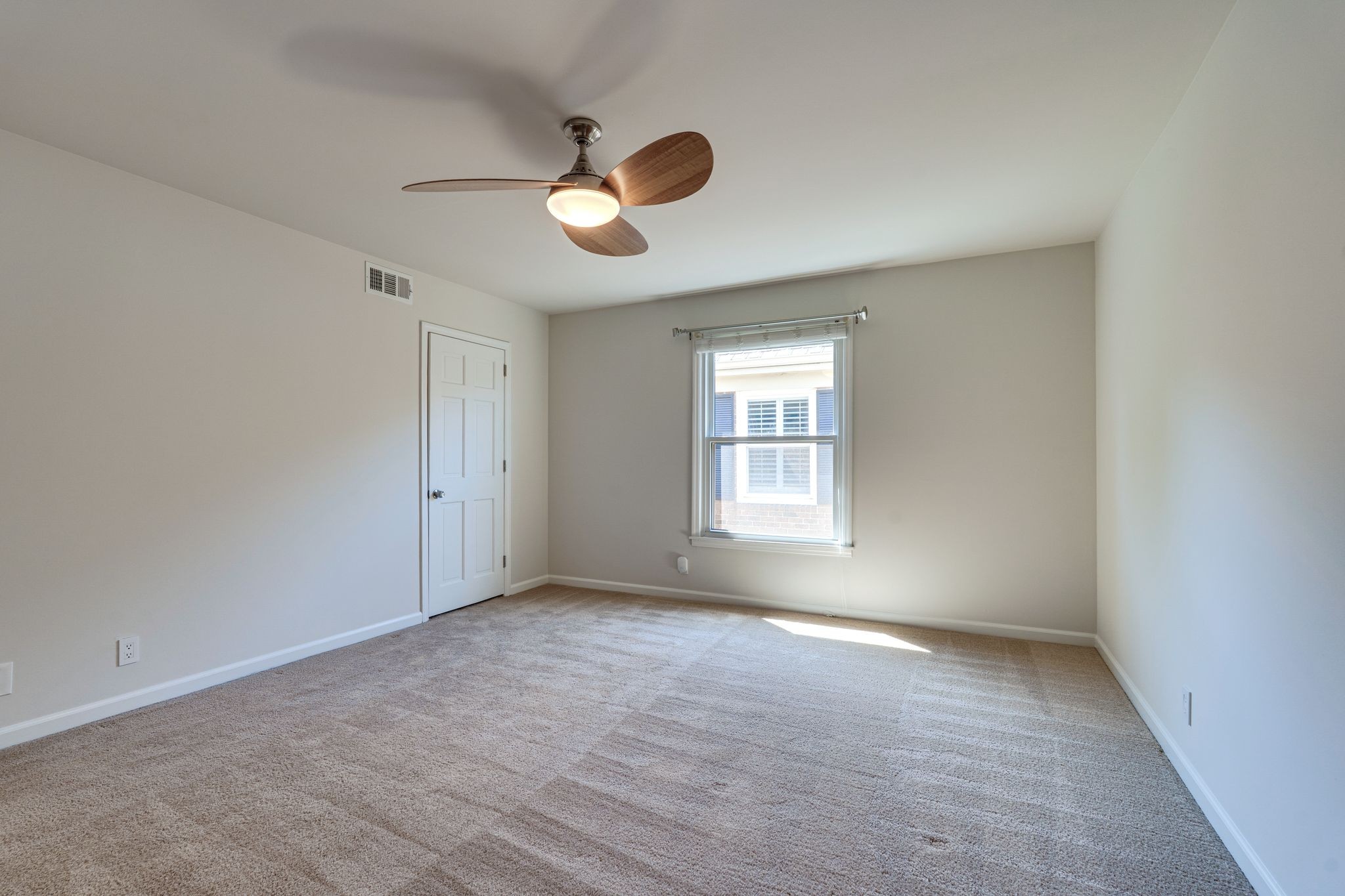 Empty carpeted bedroom with a wood-blade ceiling fan and a sunlit window.