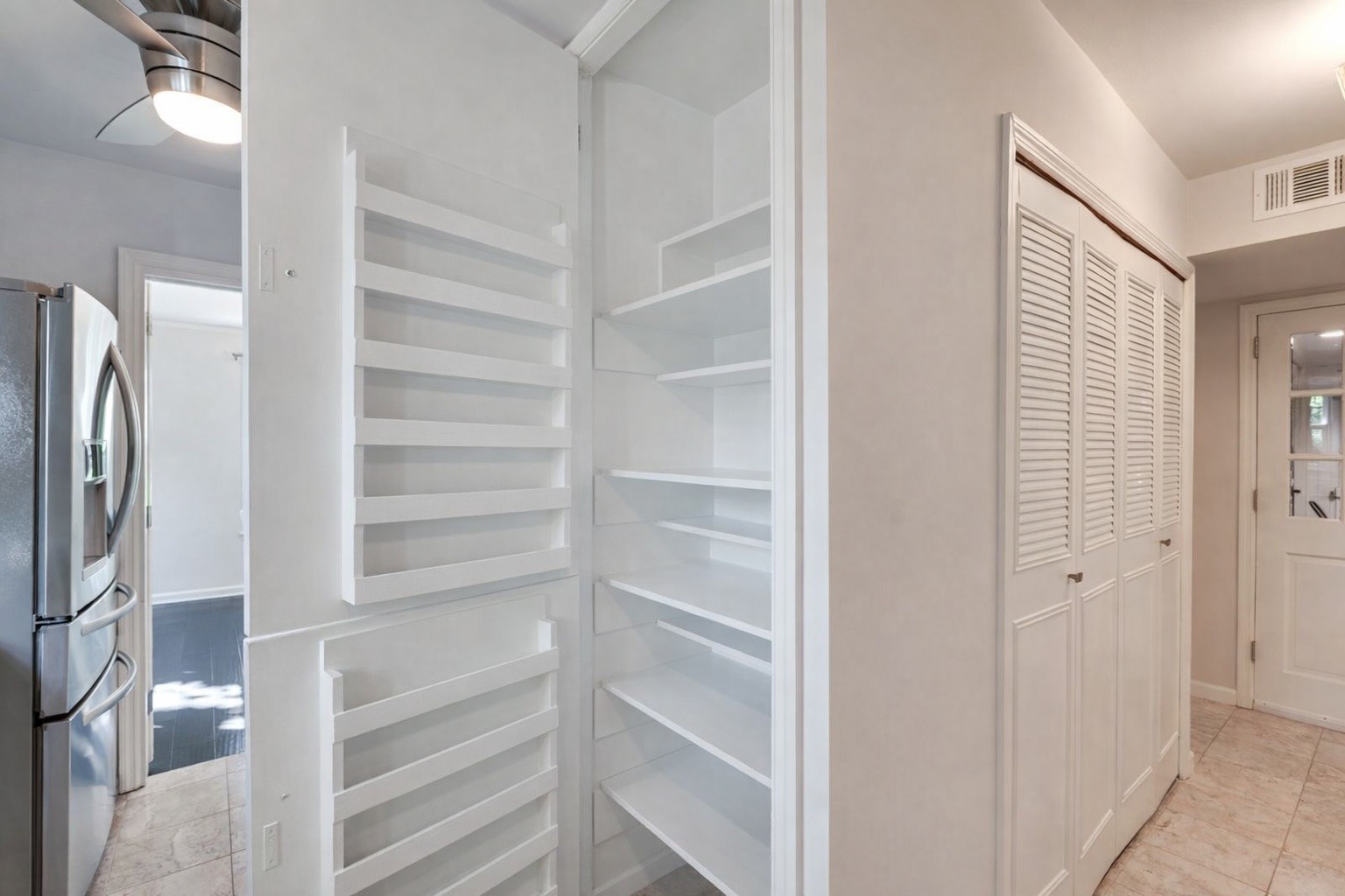 Kitchen pantry with open white shelves beside a stainless steel refrigerator and louvered cabinets.