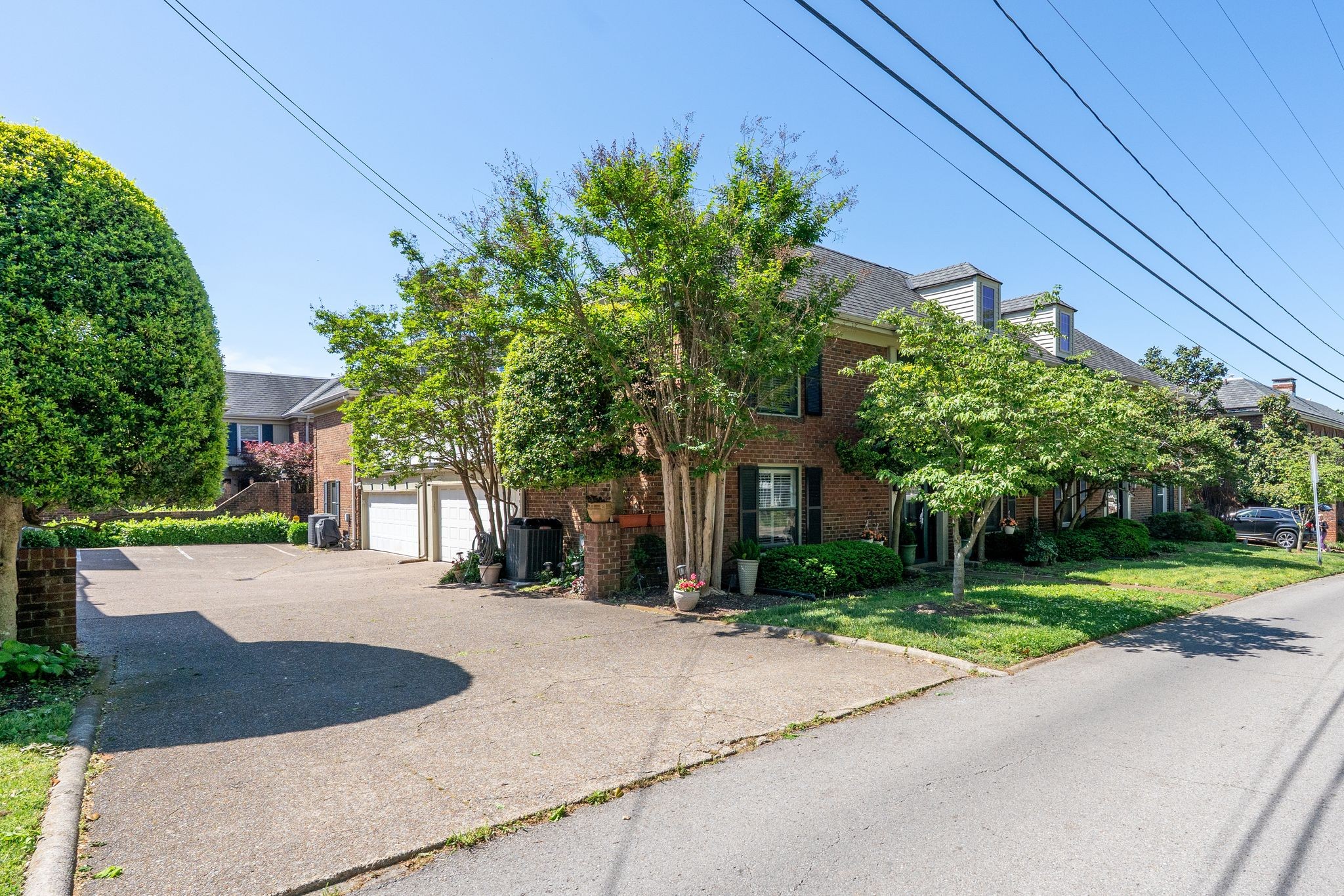 Brick townhouse partly screened by mature trees, with a paved driveway and white garage doors.