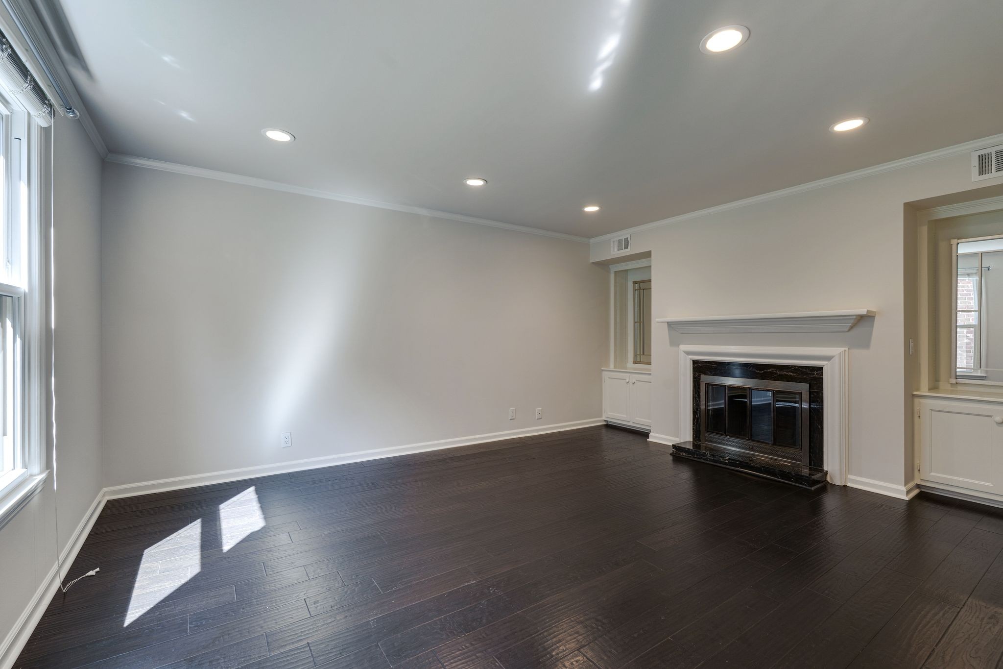 Vacant living room with dark hardwood floors, tall windows, and a black marble fireplace.