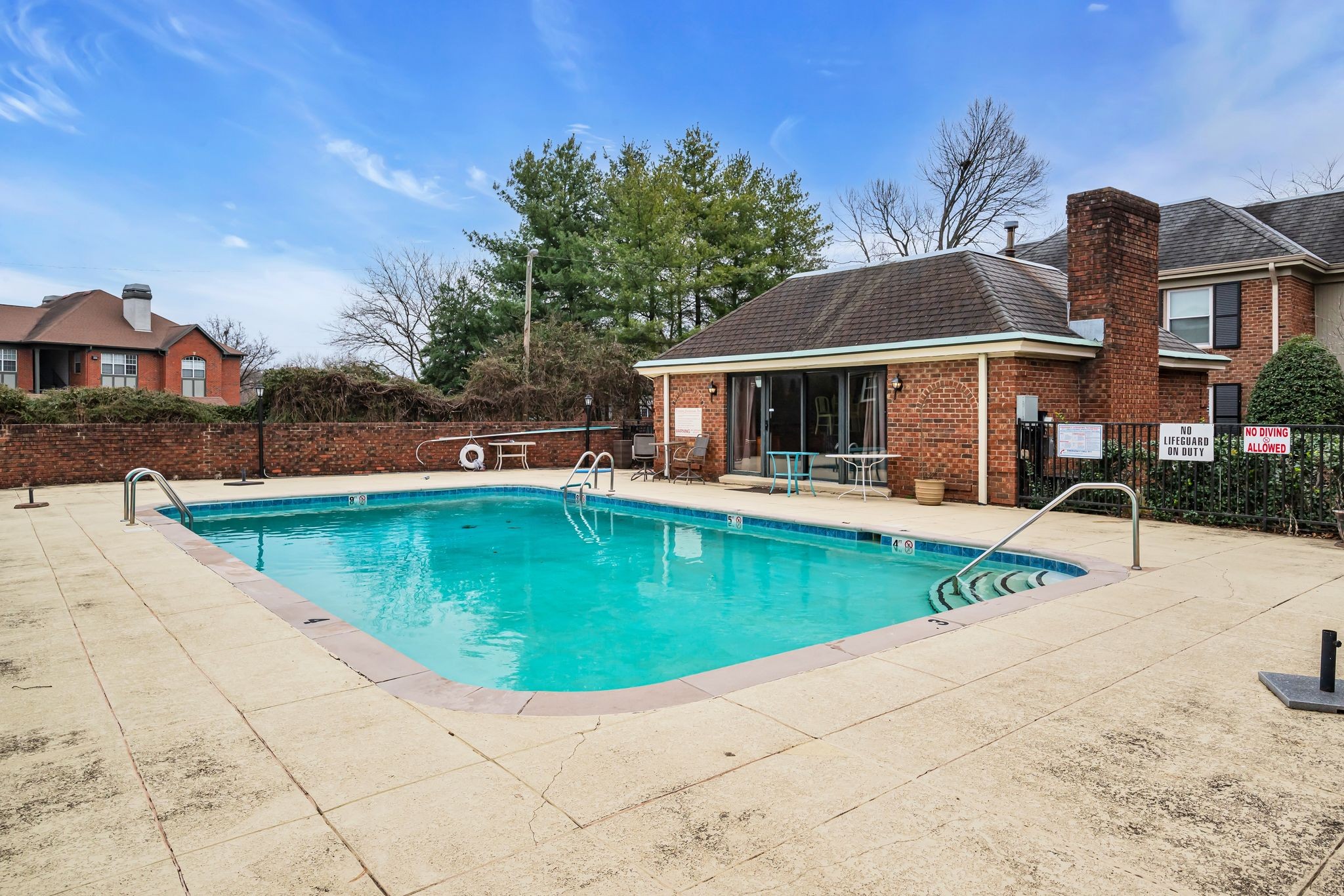 Outdoor swimming pool beside a brick pool house, with a wide paved deck and metal handrails.