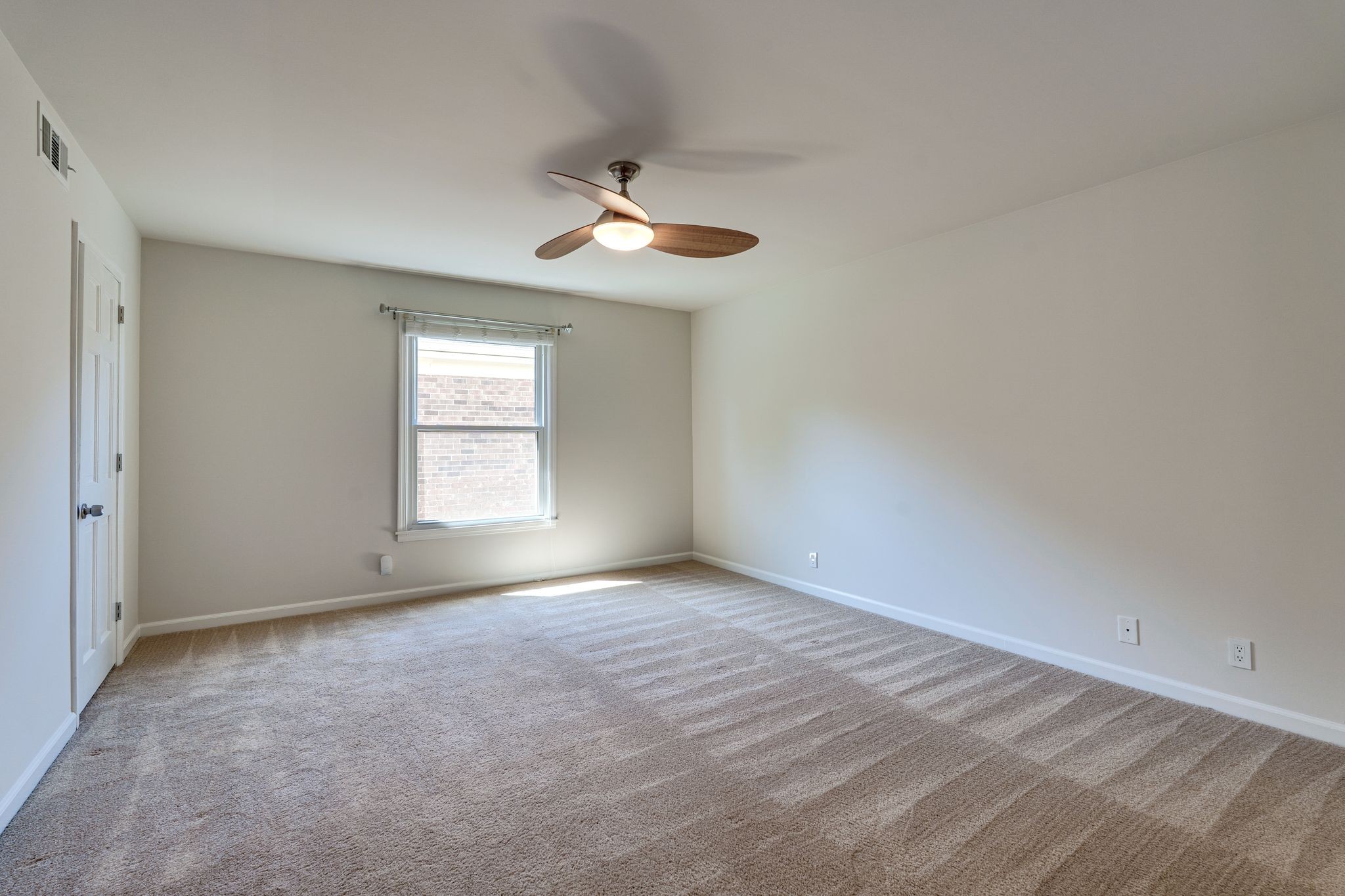 Empty bedroom with beige carpet, a window, and a wood-bladed ceiling fan.