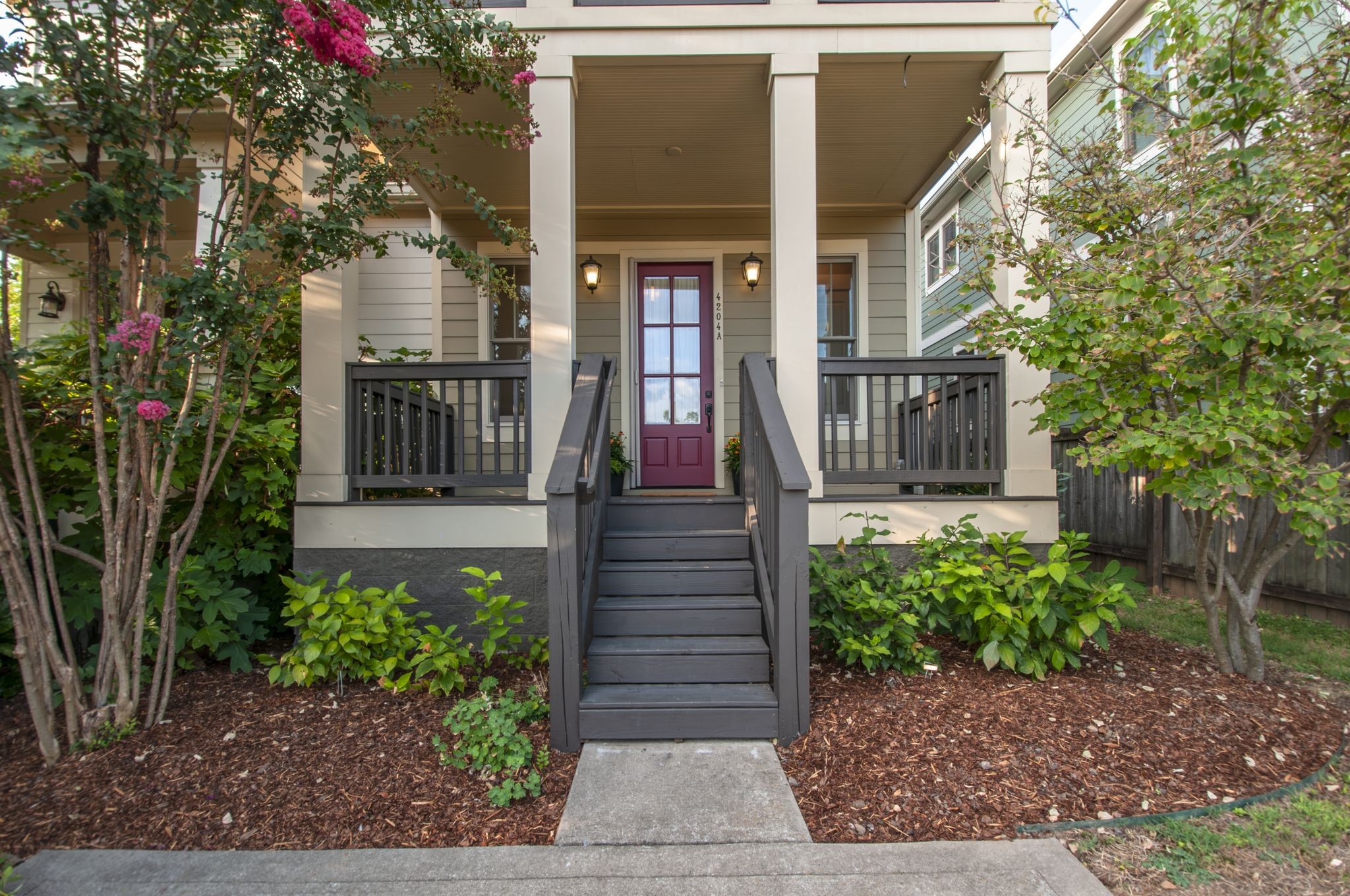 Home entrance with a covered porch, gray steps, burgundy door, and flowering shrubs.