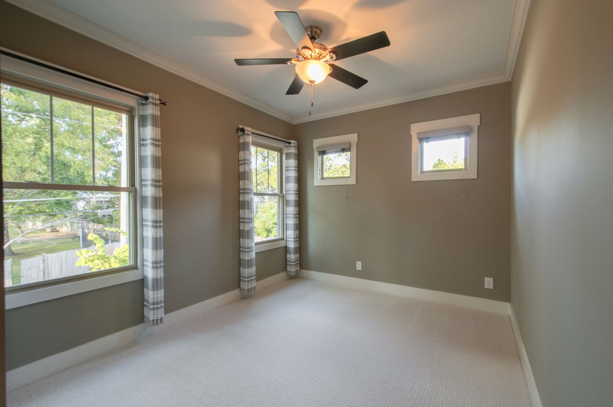 Empty bedroom with three curtained windows, light carpet, and a ceiling fan.
