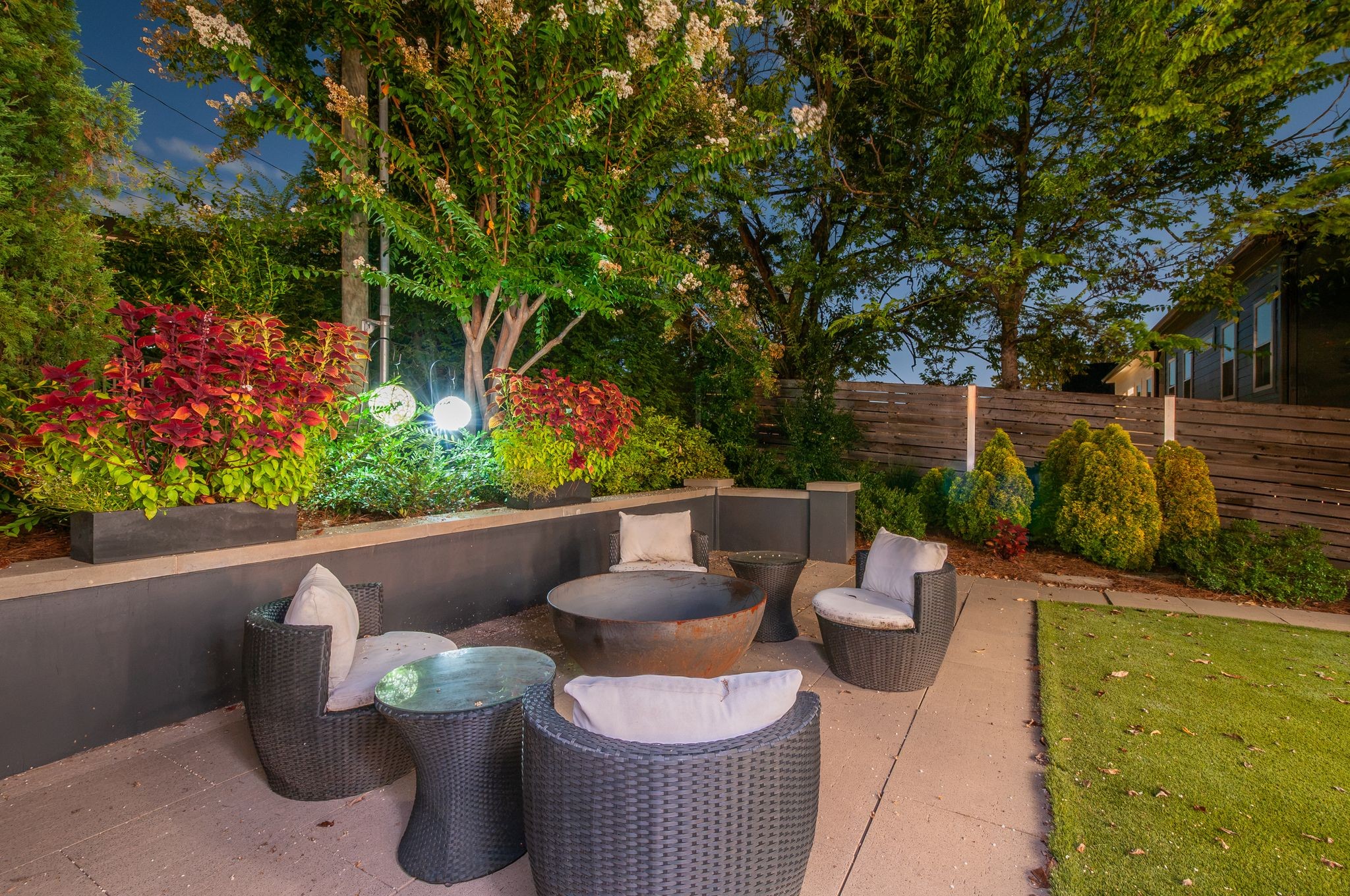 Outdoor patio with wicker seating and a round fire pit, surrounded by illuminated landscaped planters.