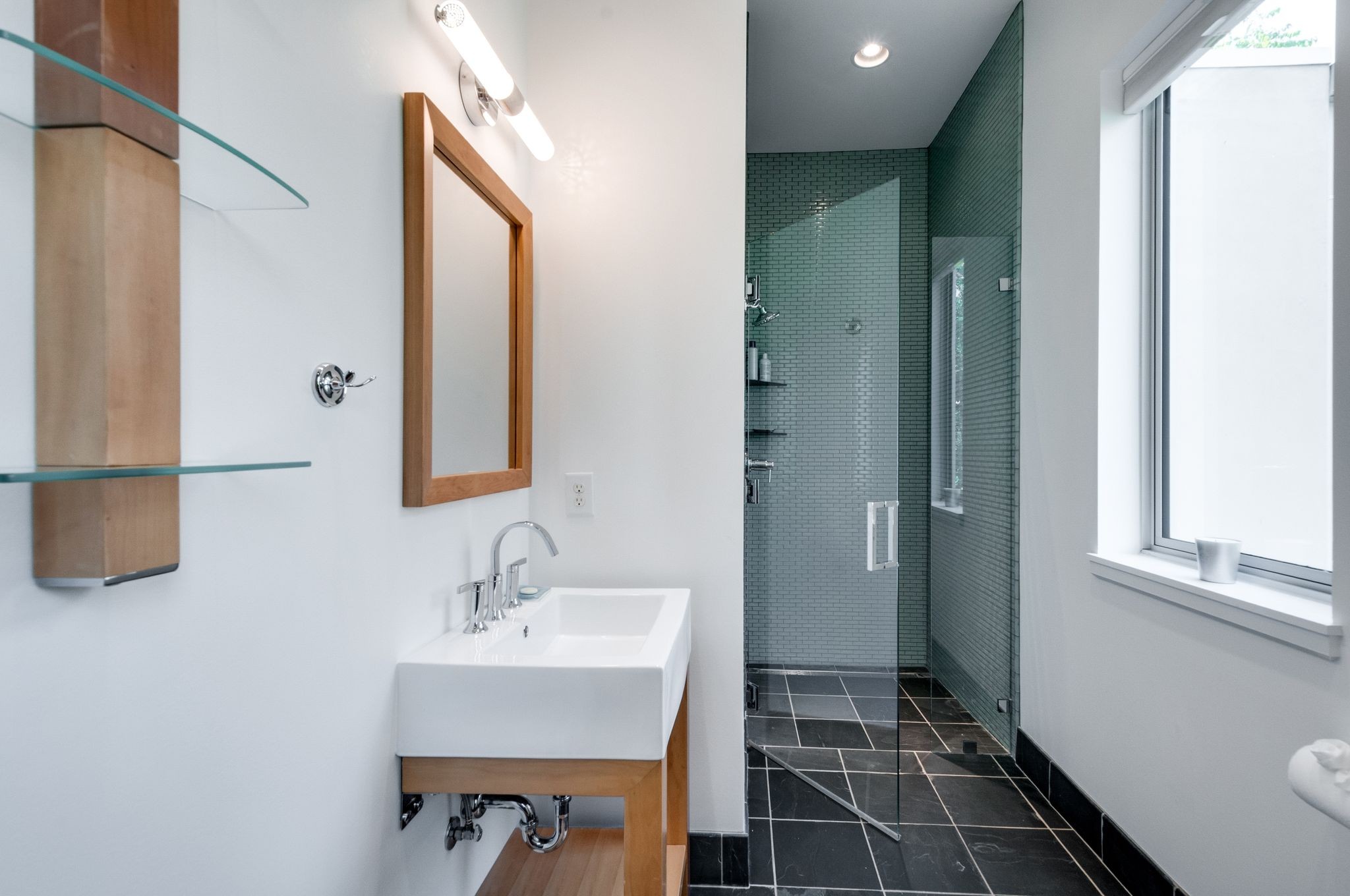 Modern bathroom with a wood vanity and glass shower lined in dark green tile.
