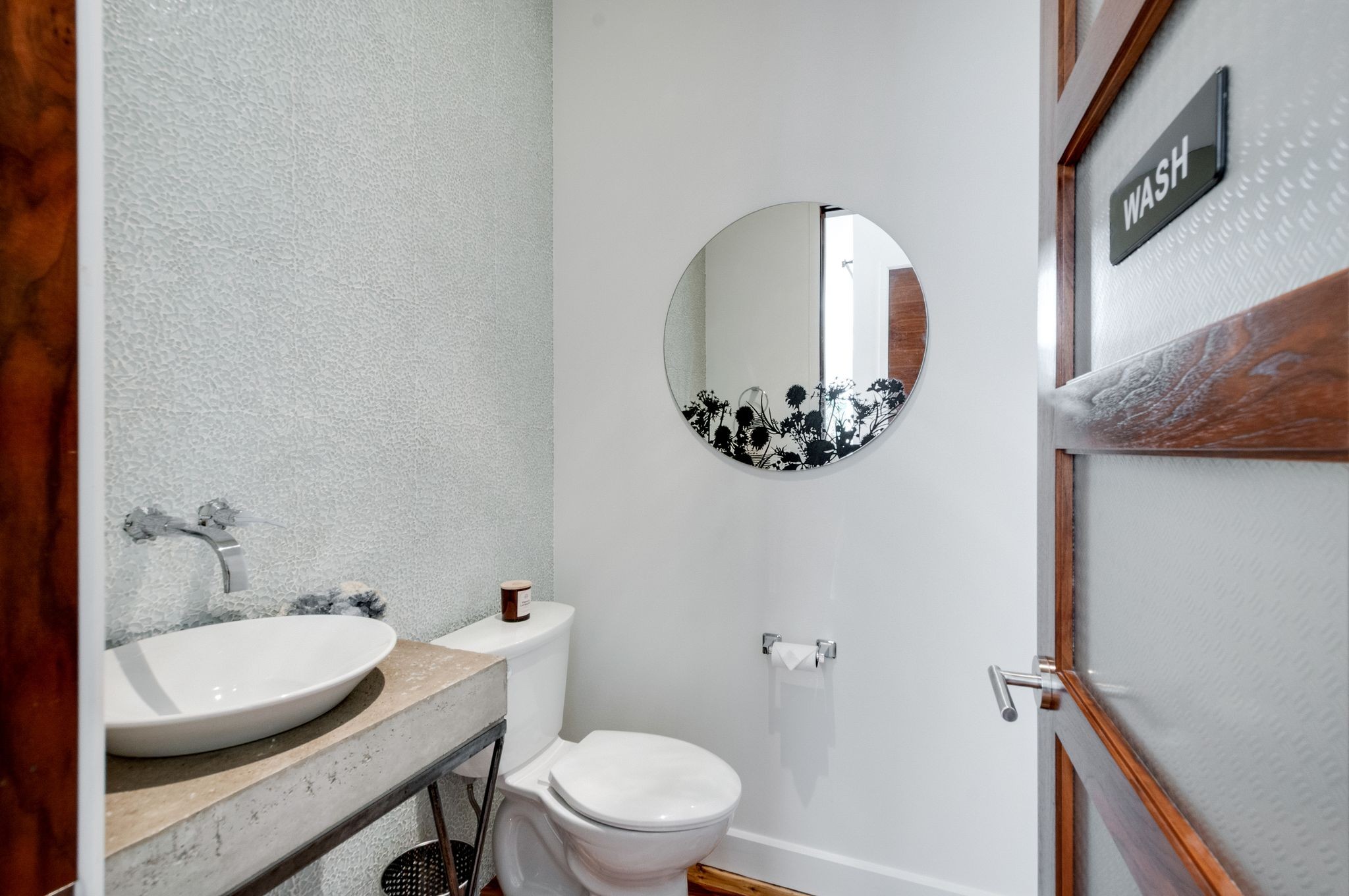 Powder room with a vessel sink, textured tile wall, round mirror, and wood-framed door marked “WASH.”