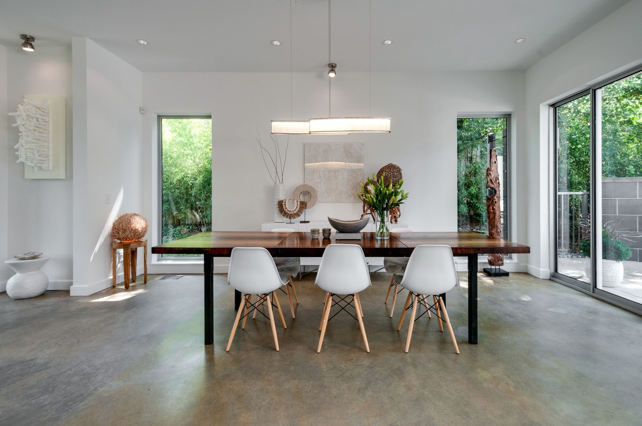 Contemporary dining room with a long wood table and white chairs beside large windows.