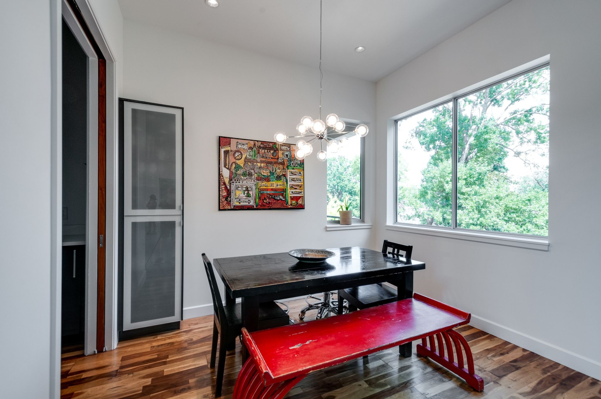Dining room with a red bench, dark wood table, globe chandelier, and large windows overlooking trees.