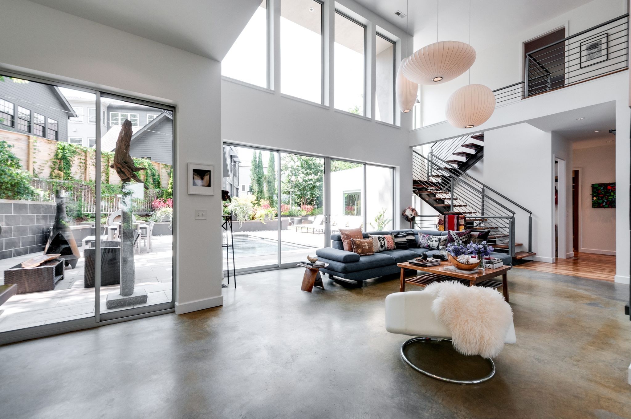 Double-height living room with a gray sectional, floating staircase, and large windows opening to a patio.